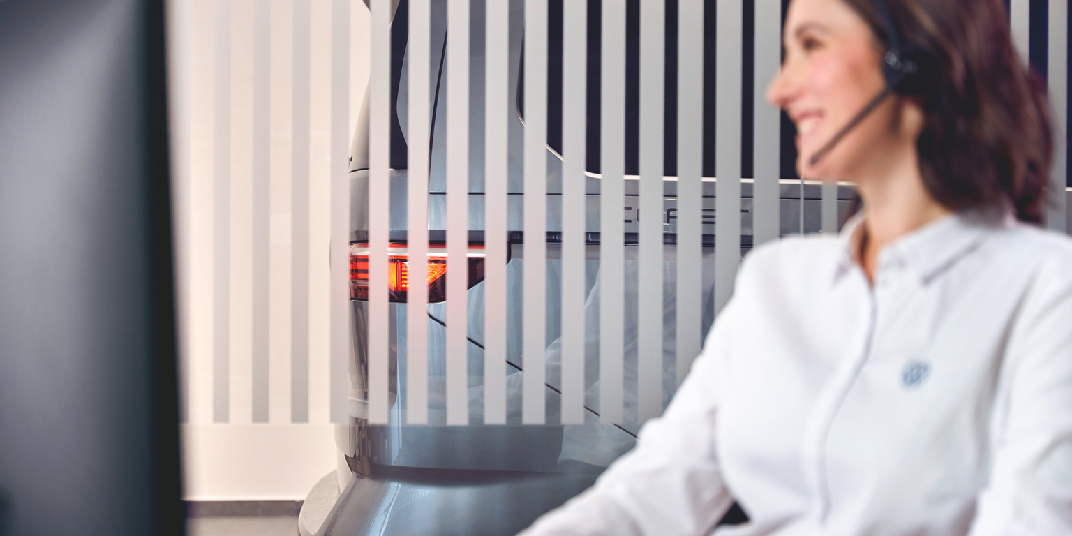 Eine Person in einem weißen Hemd sitzt vor einem Computerbildschirm. Hinter ihr steht ein graues Auto, das teilweise von vertikalen Streifen verdeckt wird. Die Rücklichter des Autos sind sichtbar.