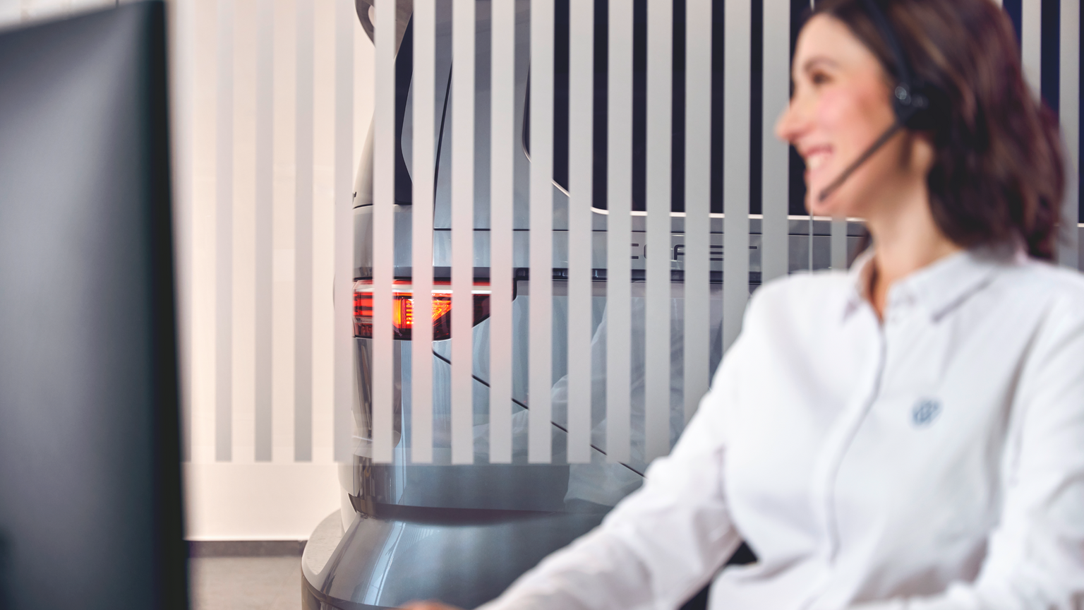 Eine Person in einem weißen Hemd sitzt vor einem Computerbildschirm. Hinter ihr steht ein graues Auto, das teilweise von vertikalen Streifen verdeckt wird. Die Rücklichter des Autos sind sichtbar.