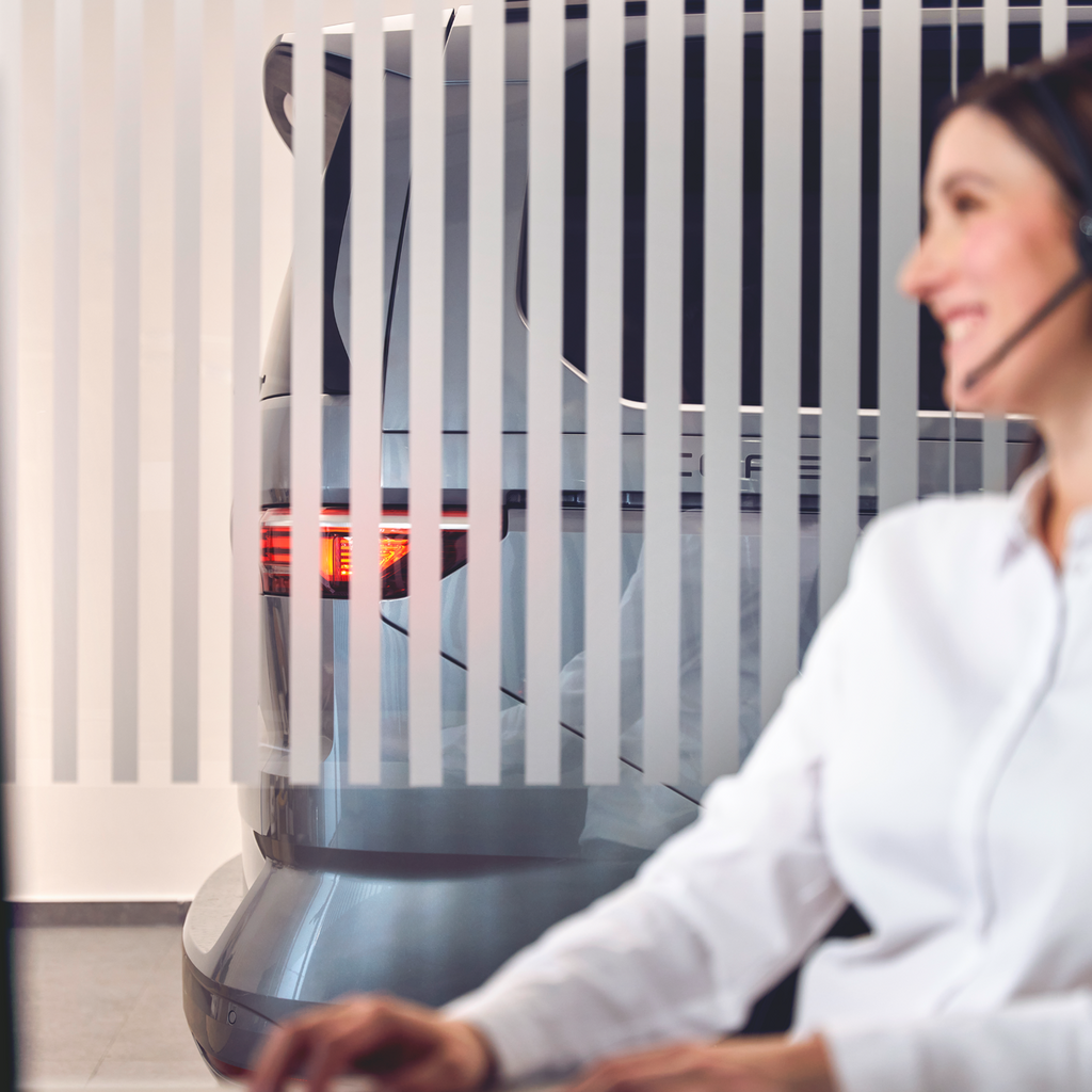 Eine Person in einem weißen Hemd sitzt vor einem Computerbildschirm. Hinter ihr steht ein graues Auto, das teilweise von vertikalen Streifen verdeckt wird. Die Rücklichter des Autos sind sichtbar.