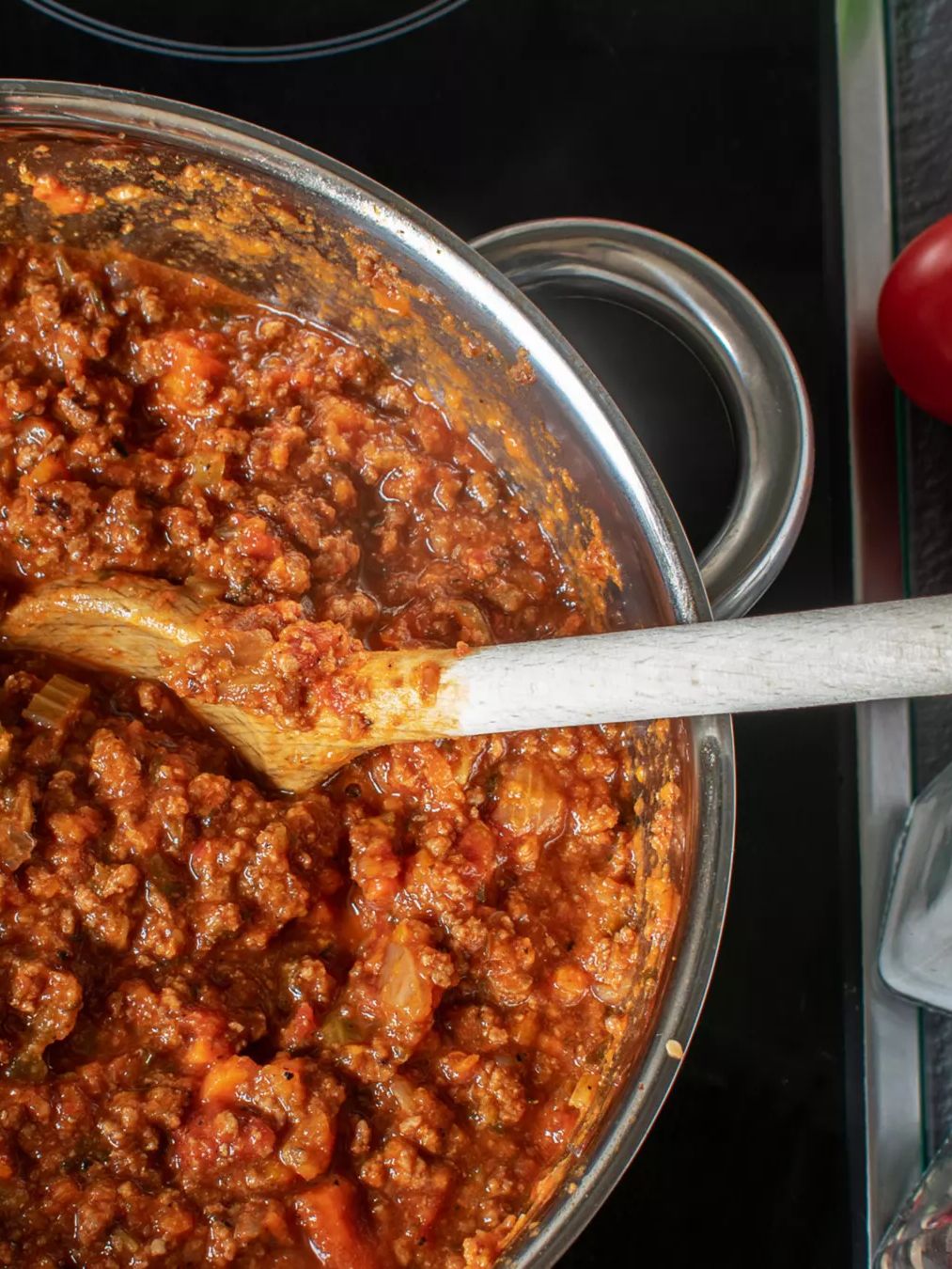 Ein großer Topf mit einer Fleischsauce, die mit einem Holzlöffel umgerührt wird, steht auf einem Herd. Daneben liegen frische Tomaten und zwei Gewürzstreuer.