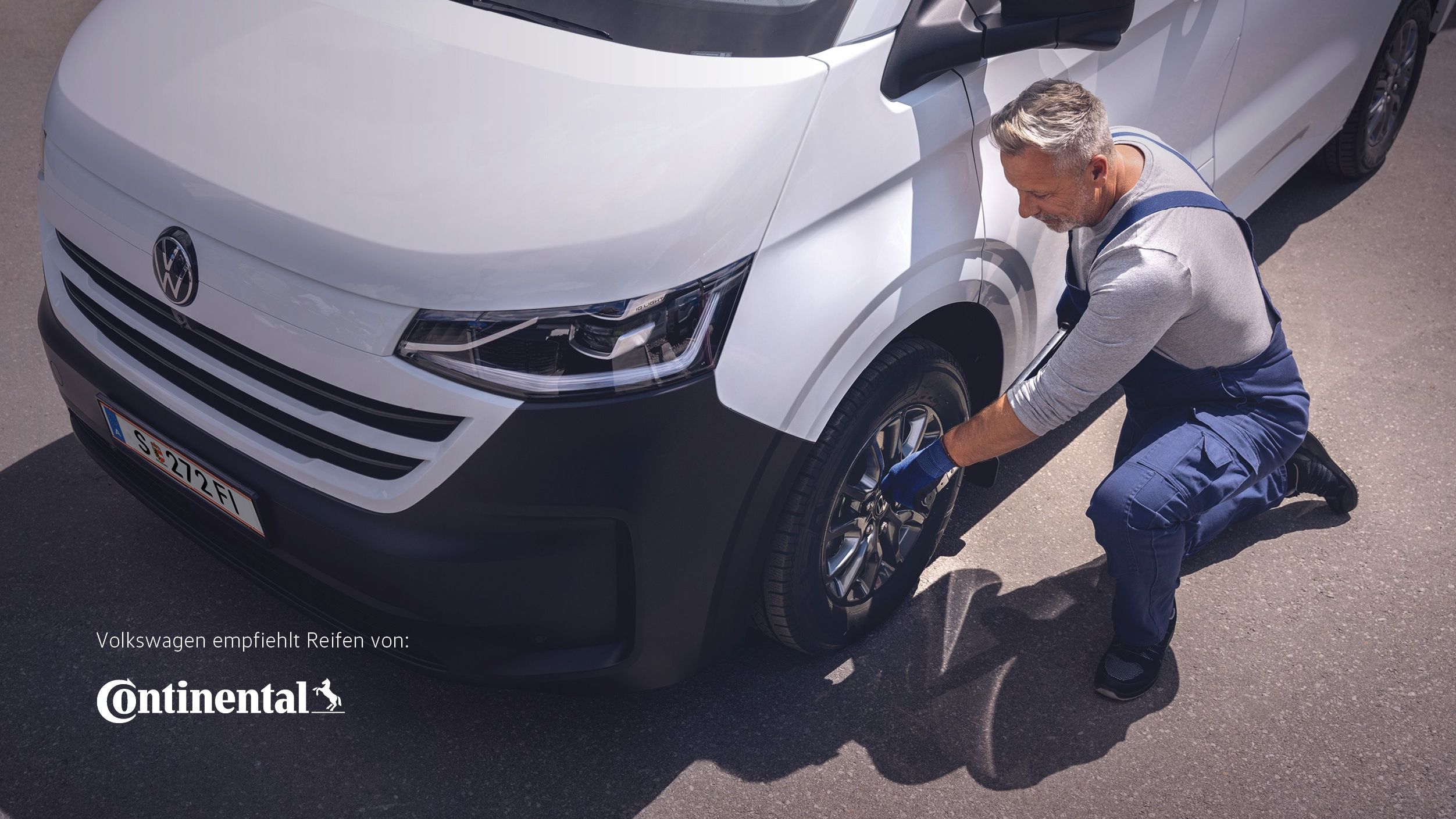 Ein Volkswagen Techniker in blauer Latzhose kniet neben einem weißen VW-Transporter und zieht mit einem Radkreuz die Radmuttern eines Vorderrads an.
