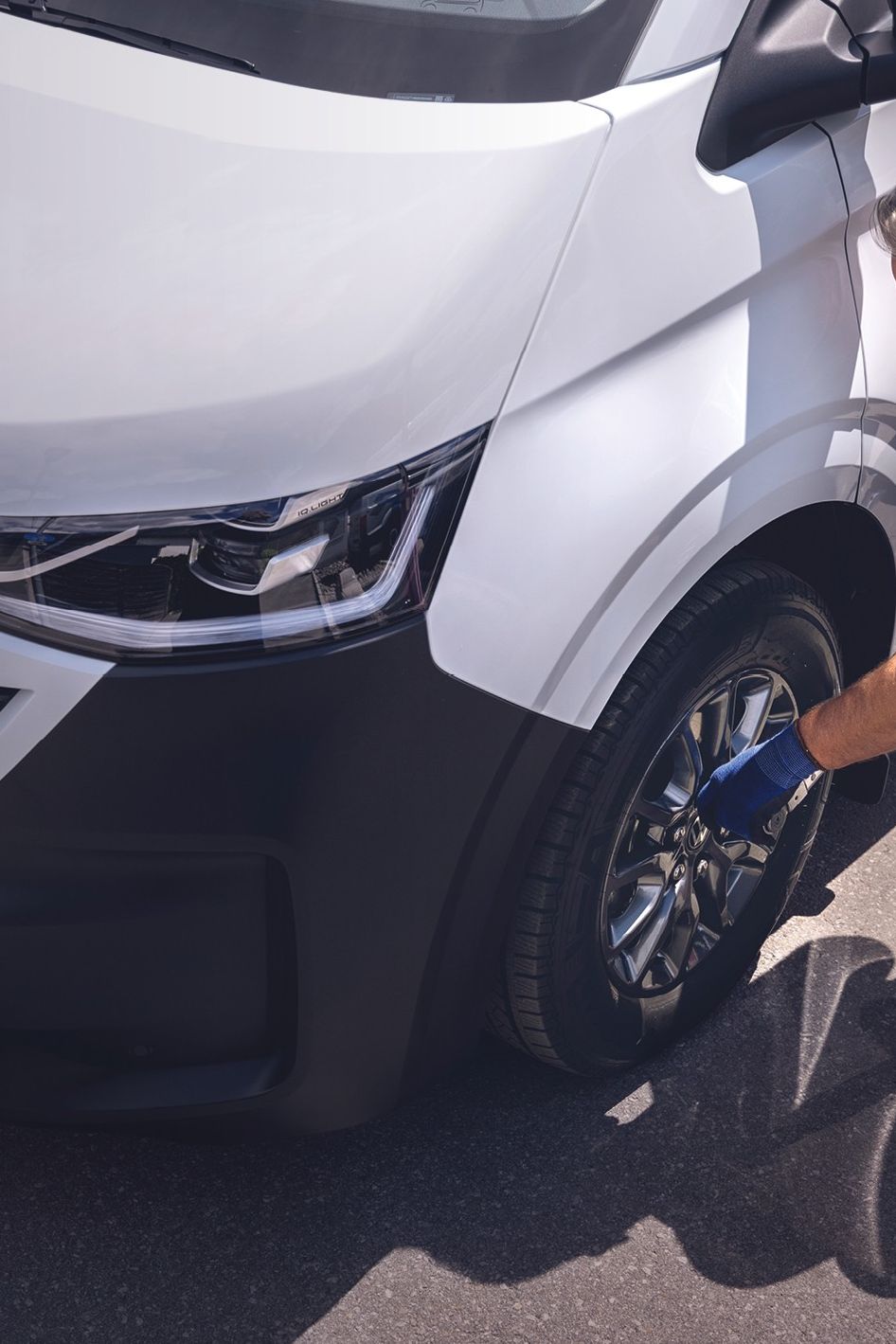 Ein Volkswagen Techniker in blauer Latzhose kniet neben einem weißen VW-Transporter und zieht mit einem Radkreuz die Radmuttern eines Vorderrads an.