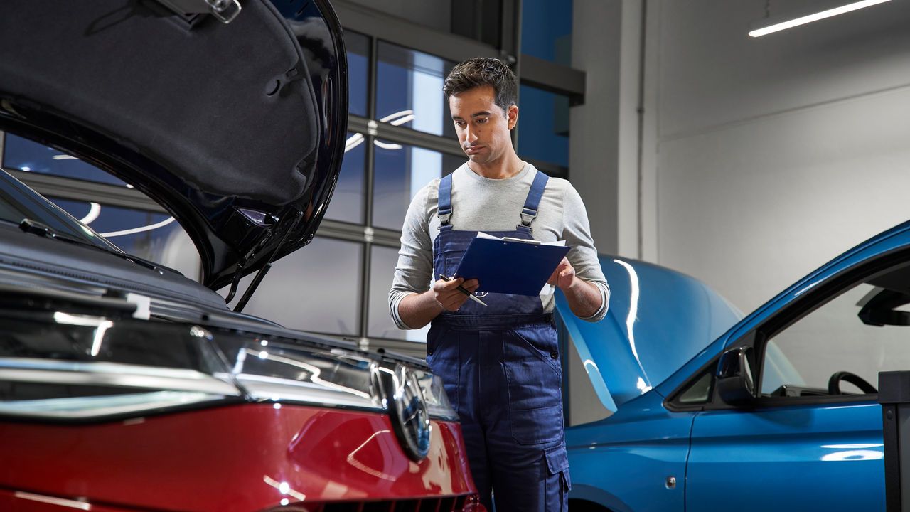 überprüft einen roten und einen blauen Volkswagen in einer Werkstatt. Die Volkswagen Werkstatt hat moderne Beleuchtung und große Fenster im Hintergrund.