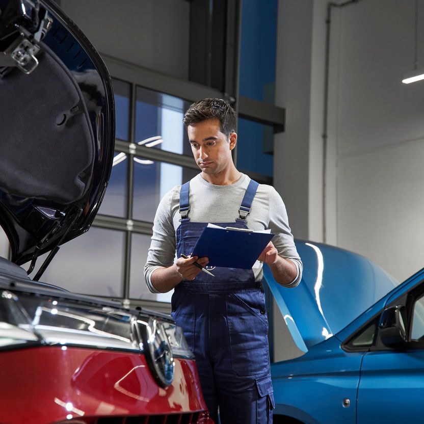 überprüft einen roten und einen blauen Volkswagen in einer Werkstatt. Die Volkswagen Werkstatt hat moderne Beleuchtung und große Fenster im Hintergrund.