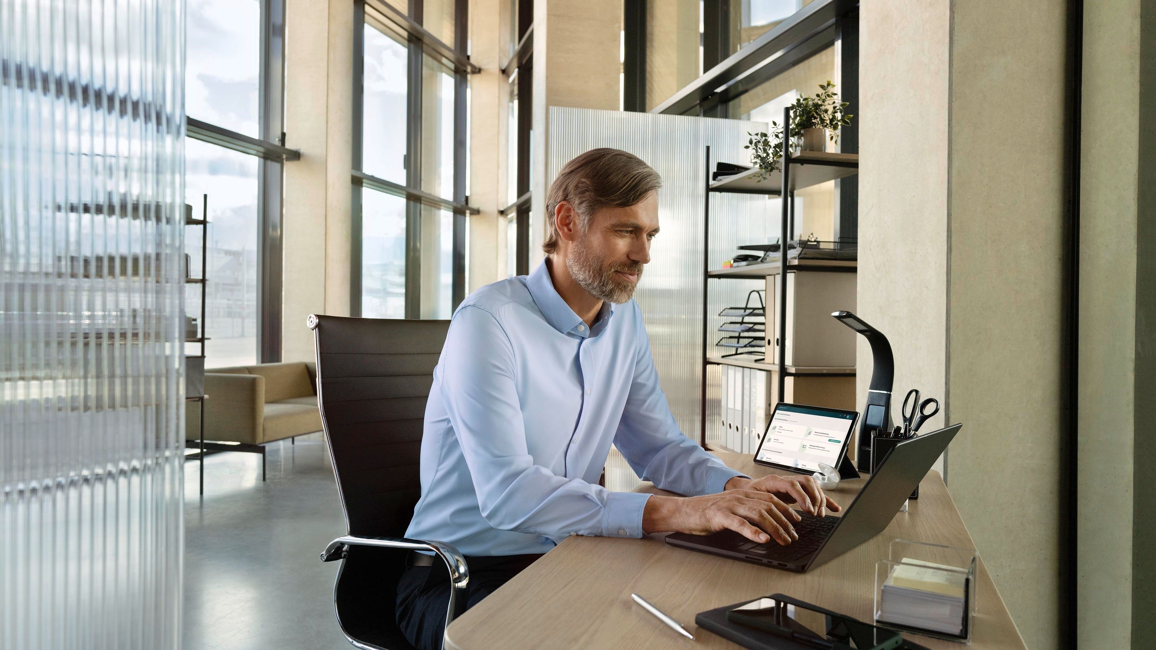 Ein Mensch sitzt an einem Schreibtisch in einem modernen Büro und arbeitet am Laptop. Auf dem Schreibtisch befinden sich weitere Arbeitsmaterialien wie Bücher, Stifte und ein Tablet. Im Hintergrund sind große Fenster und Regale sichtbar.