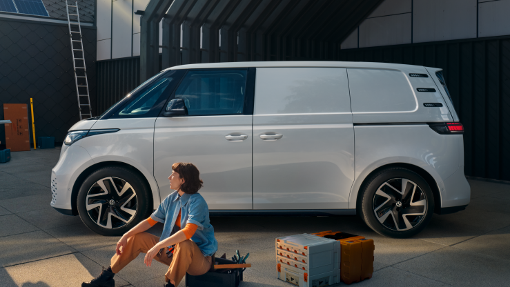 Ein weißer Volkswagen ID. Buzz Cargo vor einer Werkshalle, im Vordergrund sind ein Mann und eine Frau zu sehen