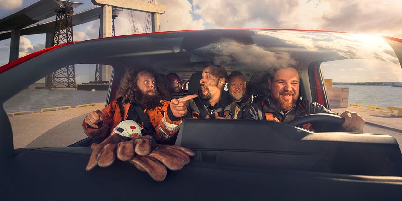 Mehrere Personen sitzen in einem Auto und tragen Sicherheitsanzüge. Im Vordergrund ist ein Kinderspielzeug in Form einer Krabbe zu sehen. Im Hintergrund sind Baustellenstrukturen und ein Wassergebiet ersichtlich.