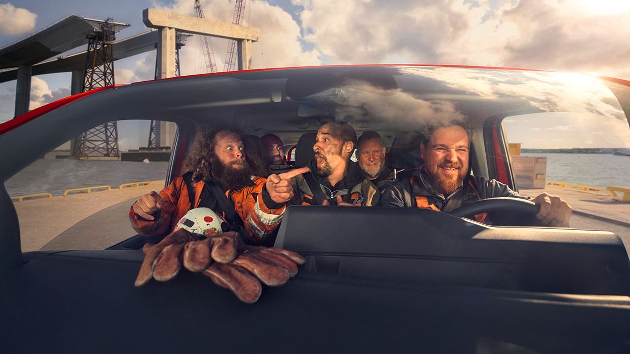 Mehrere Personen sitzen in einem Auto und tragen Sicherheitsanzüge. Im Vordergrund ist ein Kinderspielzeug in Form einer Krabbe zu sehen. Im Hintergrund sind Baustellenstrukturen und ein Wassergebiet ersichtlich.