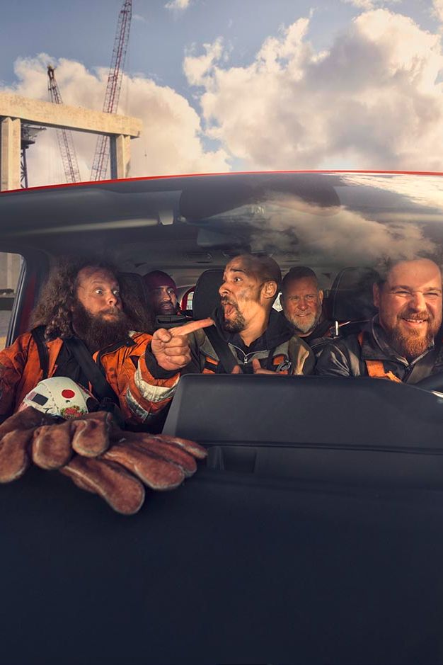 Mehrere Personen sitzen in einem Auto und tragen Sicherheitsanzüge. Im Vordergrund ist ein Kinderspielzeug in Form einer Krabbe zu sehen. Im Hintergrund sind Baustellenstrukturen und ein Wassergebiet ersichtlich.