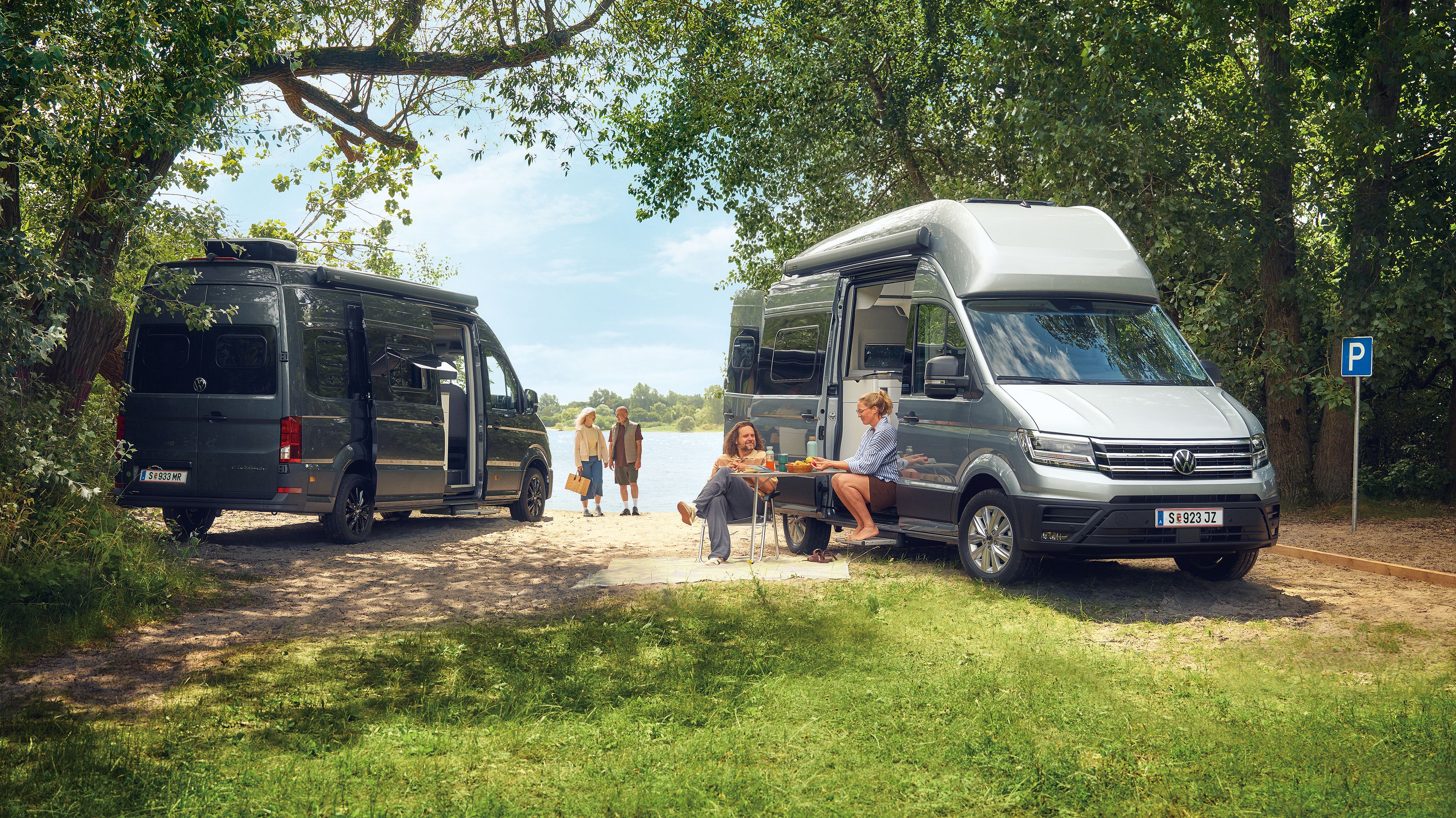 Zwei VW Grand California stehen auf einer grasbewachsenen Fläche in der Nähe eines Sees. Es gibt mehrere Personen; eine sitzt an einem Picknicktisch und zwei weitere gehen auf das Wasser zu. Die Szene ist von Bäumen und Grün umgeben.