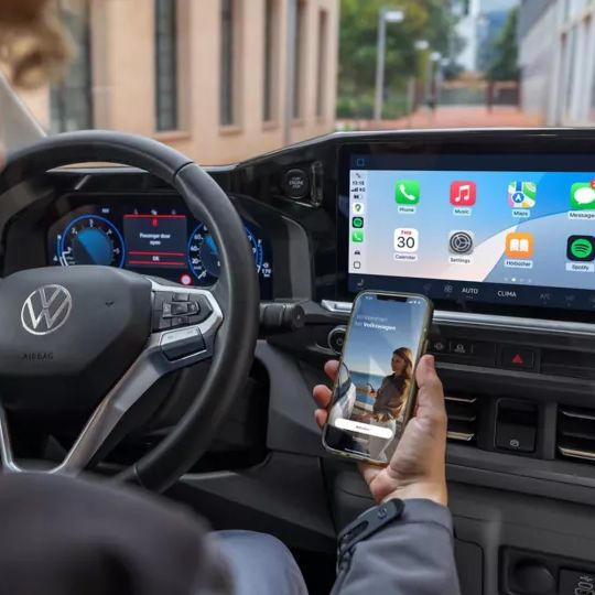 Eine Person sitzt in einem Auto und hält ein Smartphone in der Hand, während auf dem großen Display des Autos verschiedene Apps angezeigt werden. Das Lenkrad zeigt das VW-Logo.