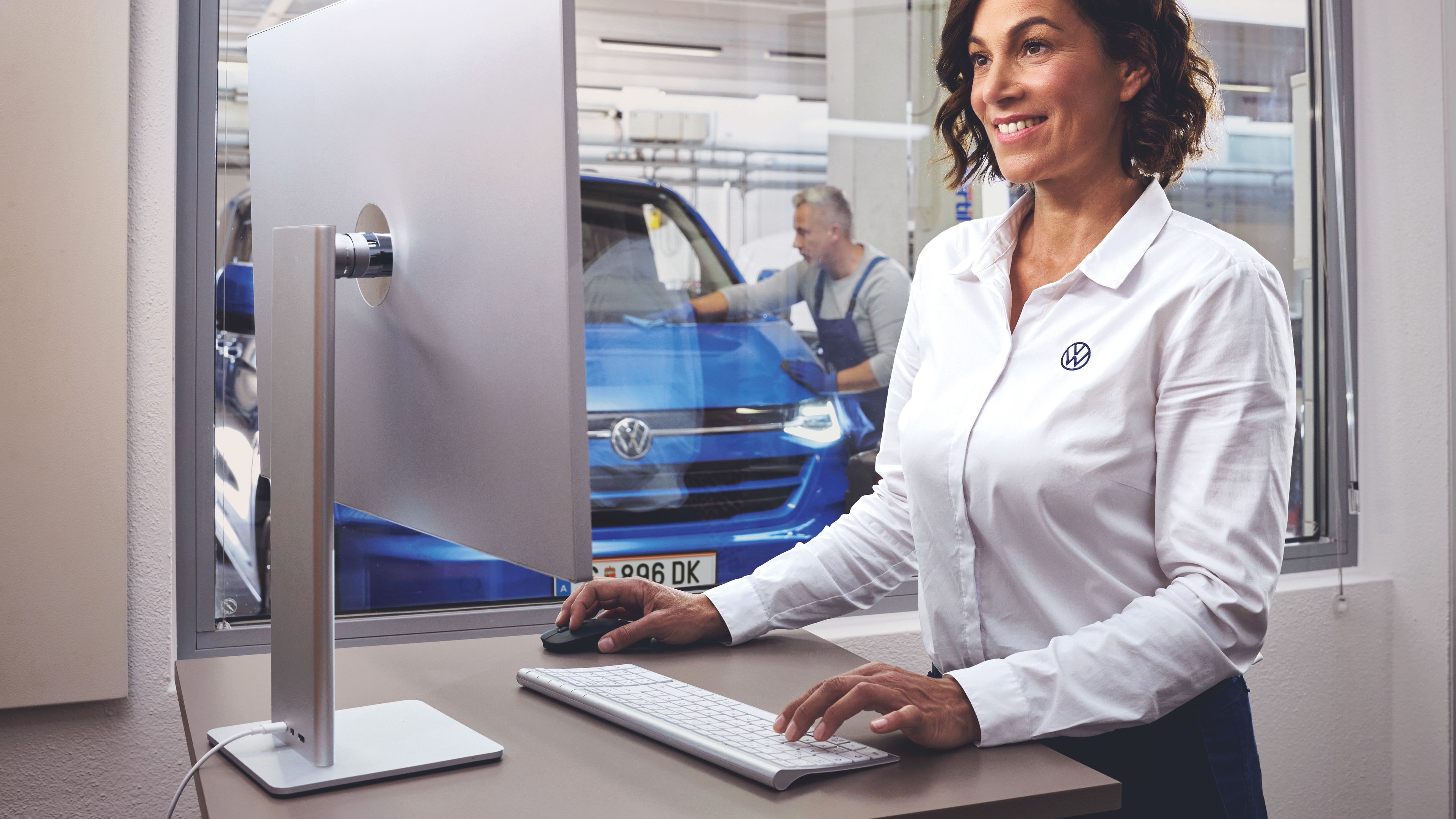 Eine Volkswagen Service MItarbeiterin tippt auf einer Tastatur und blickt auf einen Bildschirm. Im Hintergrund ist ein blauer Volkswagen Transporter in einer Werkstatt zu sehen.