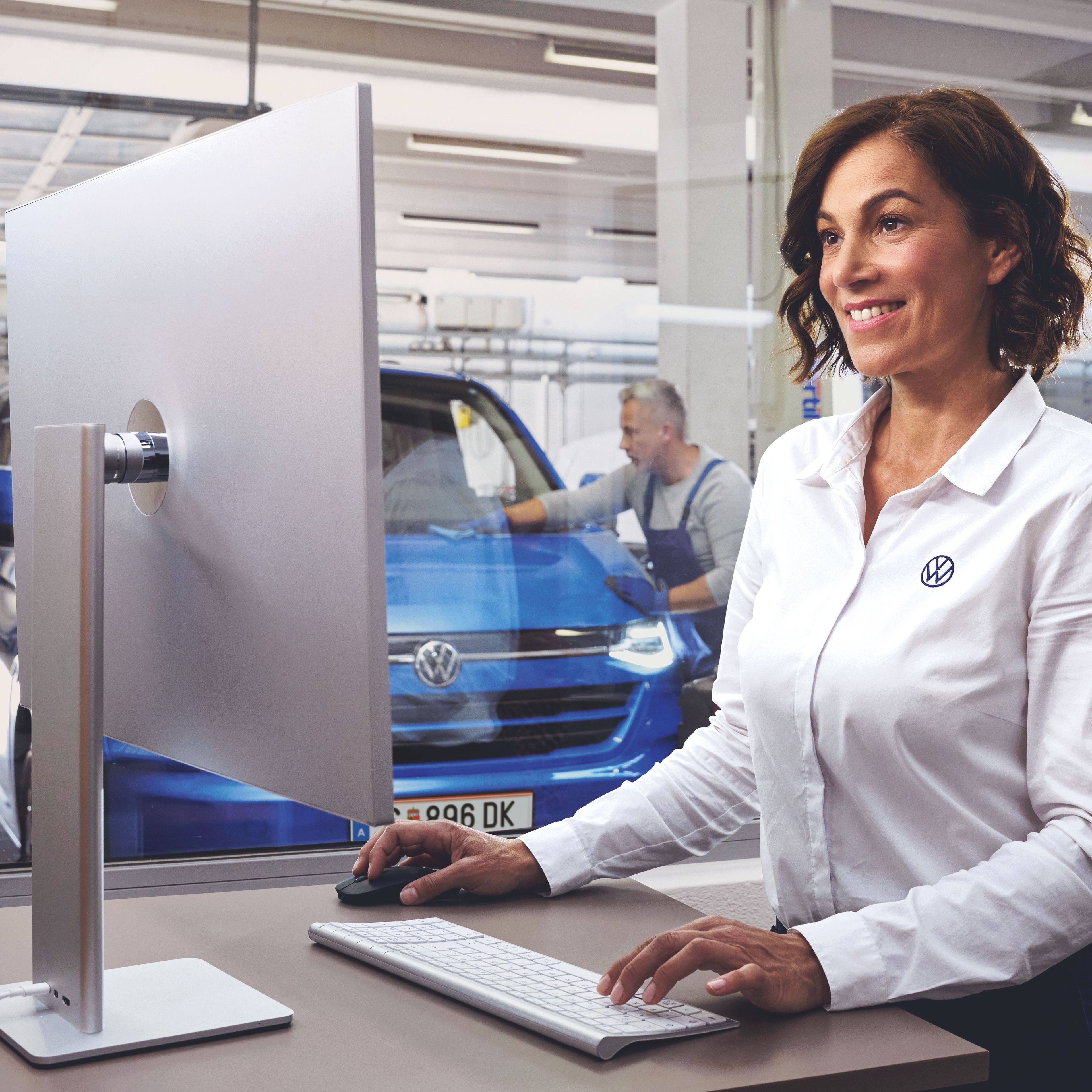 Eine Volkswagen Service MItarbeiterin tippt auf einer Tastatur und blickt auf einen Bildschirm. Im Hintergrund ist ein blauer Volkswagen Transporter in einer Werkstatt zu sehen.