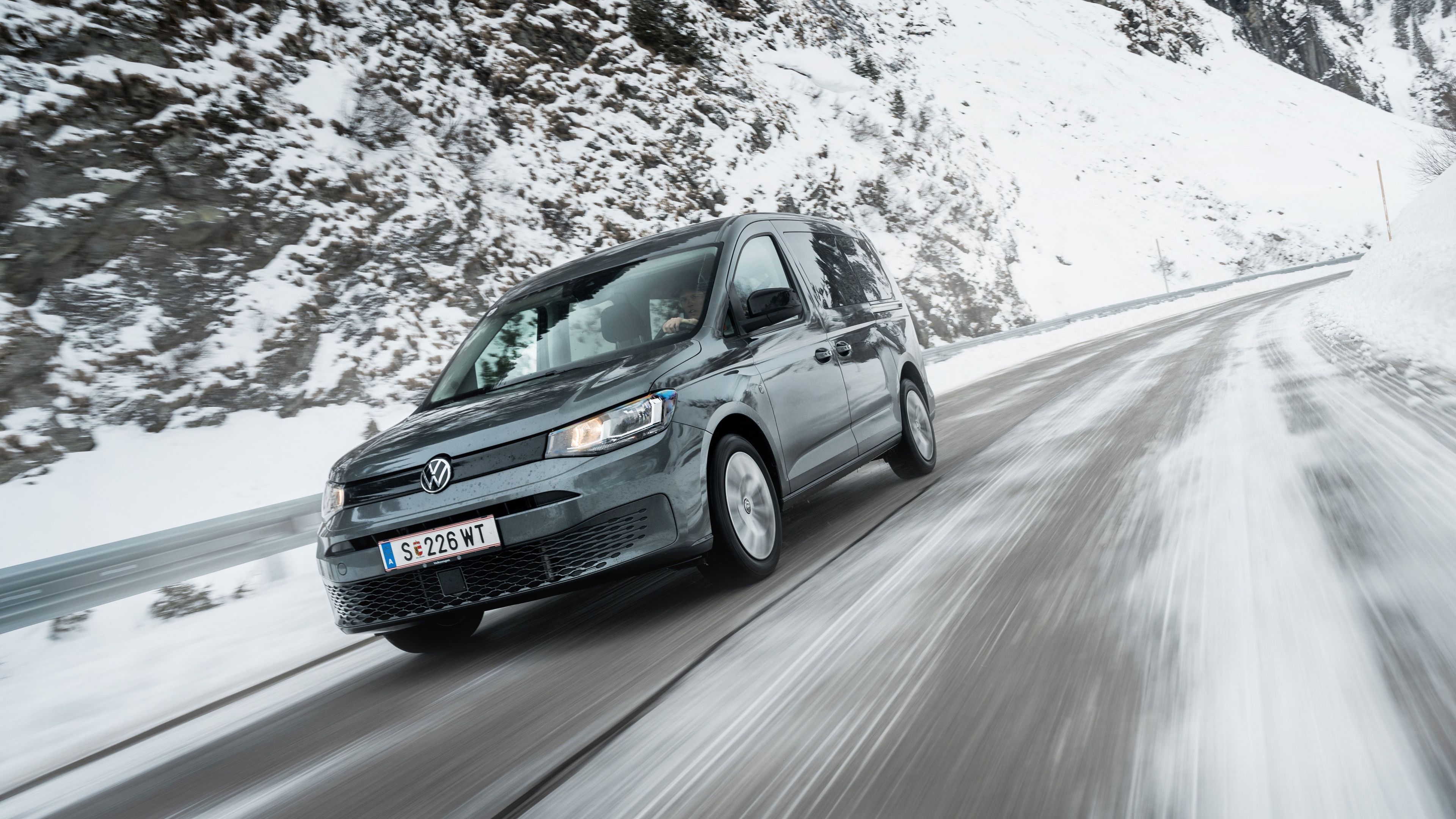 Grauer VW-Kastenwagen fährt auf einer nassen, teils verschneiten Bergstraße. Rechts und links der Straße liegen hohe Schneewände und verschneite Hänge, der Himmel ist bedeckt. Die Bewegung des Fahrzeugs wird durch verwischte Spuren auf der Fahrbahn betont.