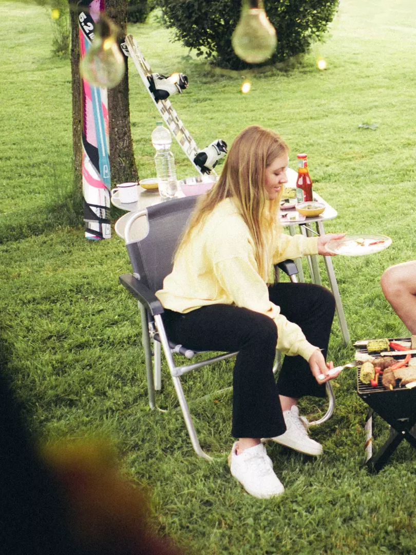 Zwei Menschen sitzen auf Campingstühlen und grillen auf einer Wiese. Sie haben Teller mit Essen und führen ein Gespräch. Im Hintergrund steht ein VW California.