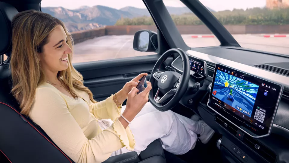 Eine Frau sitzt in einem Auto und spielt ein Videospiel auf dem eingebauten Bildschirm des Armaturenbretts. Ein Controller ist in ihren Händen zu sehen. Im Hintergrund sind Berge und eine Straße.