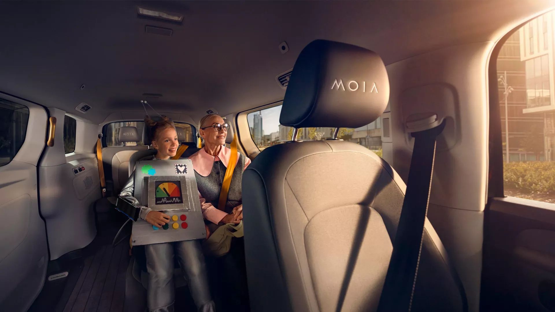Ein Kind sitzt auf dem Rücksitz eines Autos und spielt auf einem Tablet, während ein Erwachsener neben ihm sitzt. Die Sitze haben einen schwarzen Bezug mit dem Logo "MOIA". Das Auto ist geräumig und von Tageslicht durchflutet.