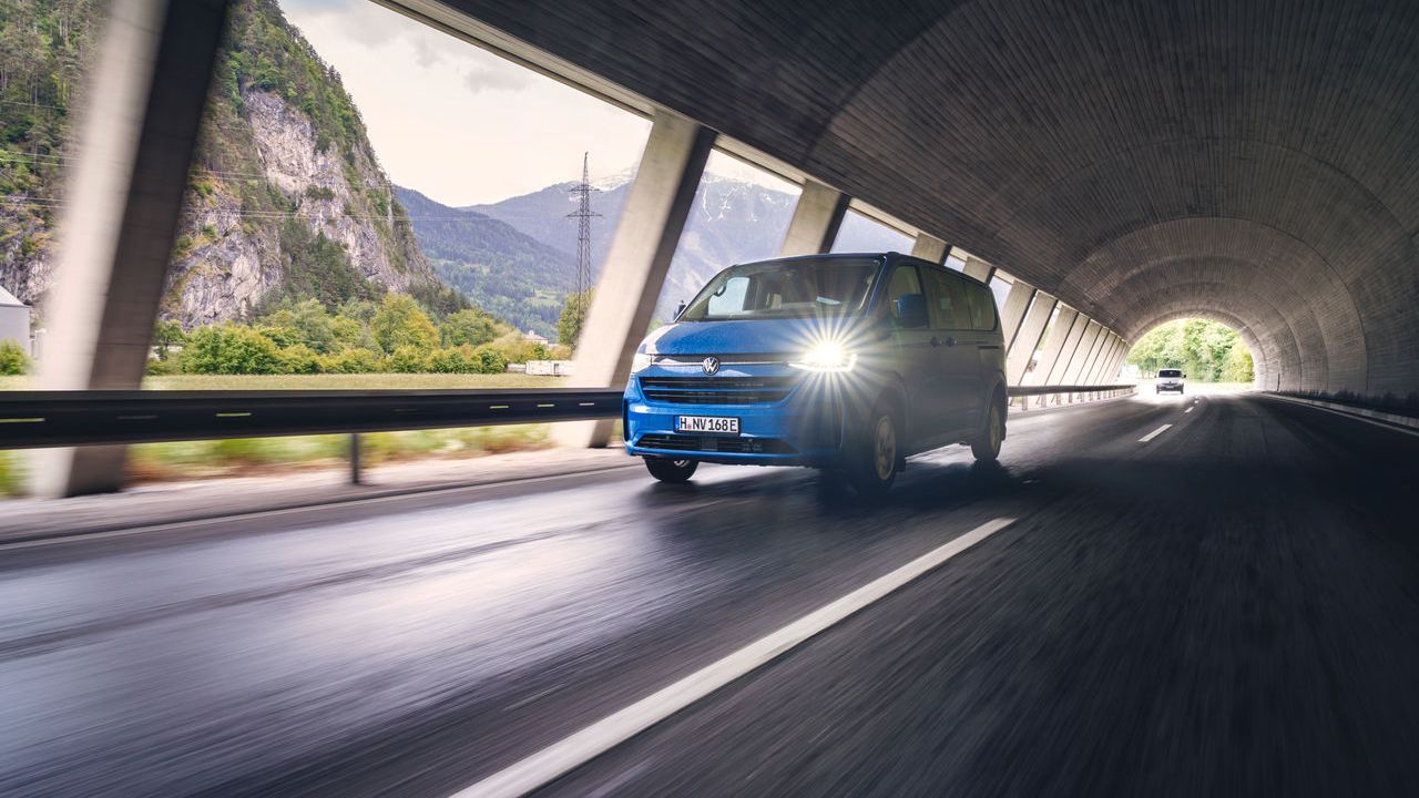 Ein blauer Volkswagen Caravelle fährt mit eingeschaltetem Licht durch einen Tunnel auf einer nassen Fahrbahn. Durch die Tunnelöffnungen sind grüne Hügel und Berge im Hintergrund zu sehen. 