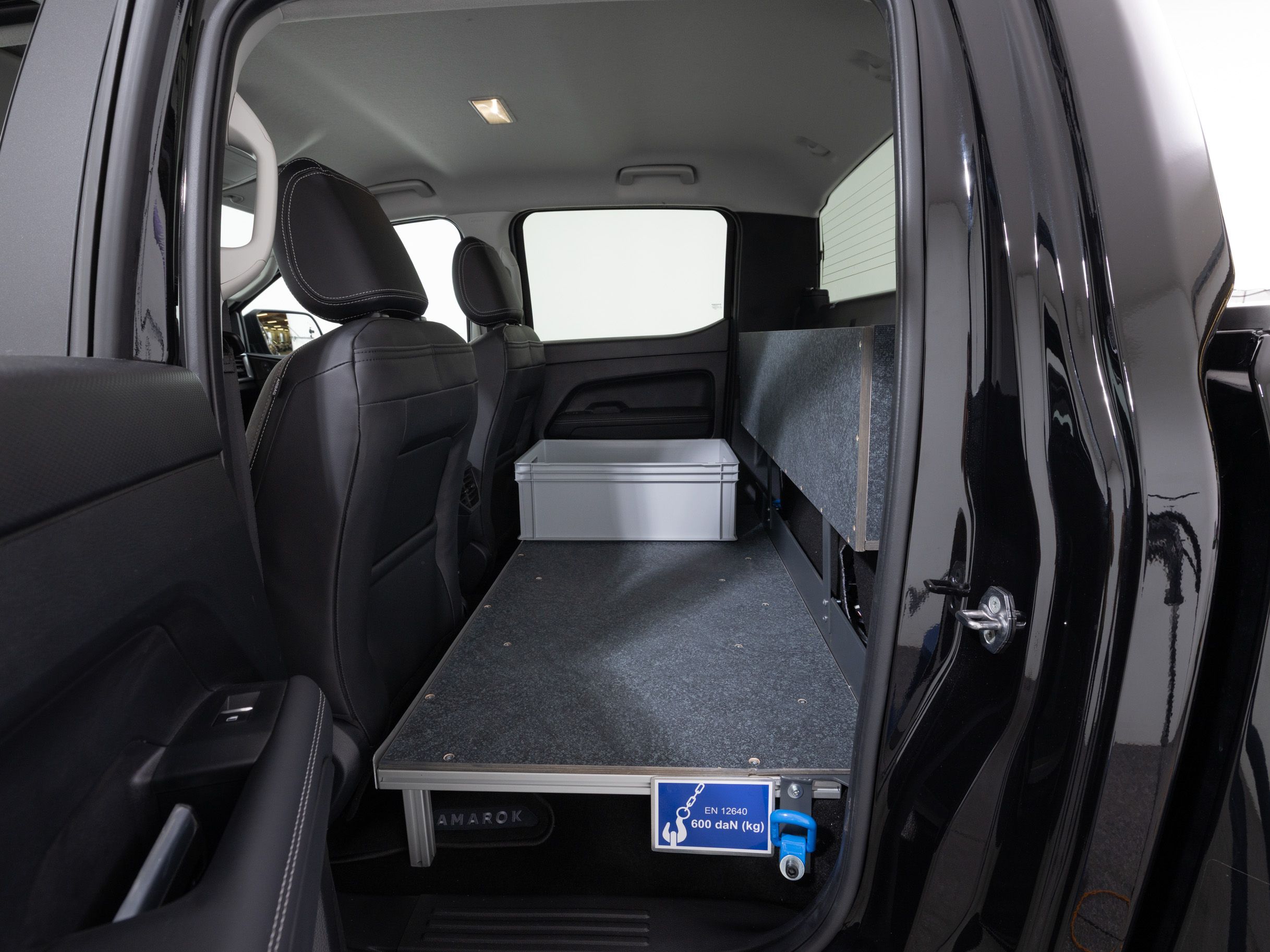 Einge Innenansicht eines Fahrzeugs zeigt den Laderaum mit einer schwarzen Plattform und einer silbernen Box. Die Rücksitze sind aus schwarzem Leder, und es gibt ein Schild an der Plattform mit einem Symbol, das auf Gebärdensprachen hinweist.