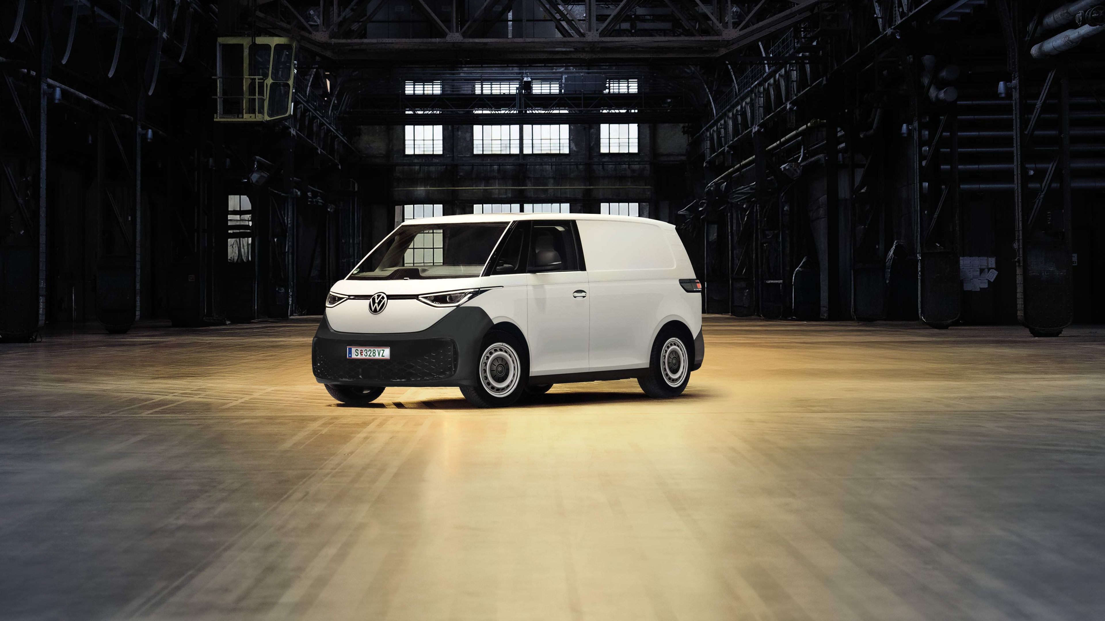 Ein weißer Lieferwagen steht allein in einer großen, dunklen Industriehalle. Die Umgebung ist leer und weist Metallstrukturen sowie Fenster im Hintergrund auf. Ein einzelner Scheinwerfer beleuchtet den Lieferwagen.