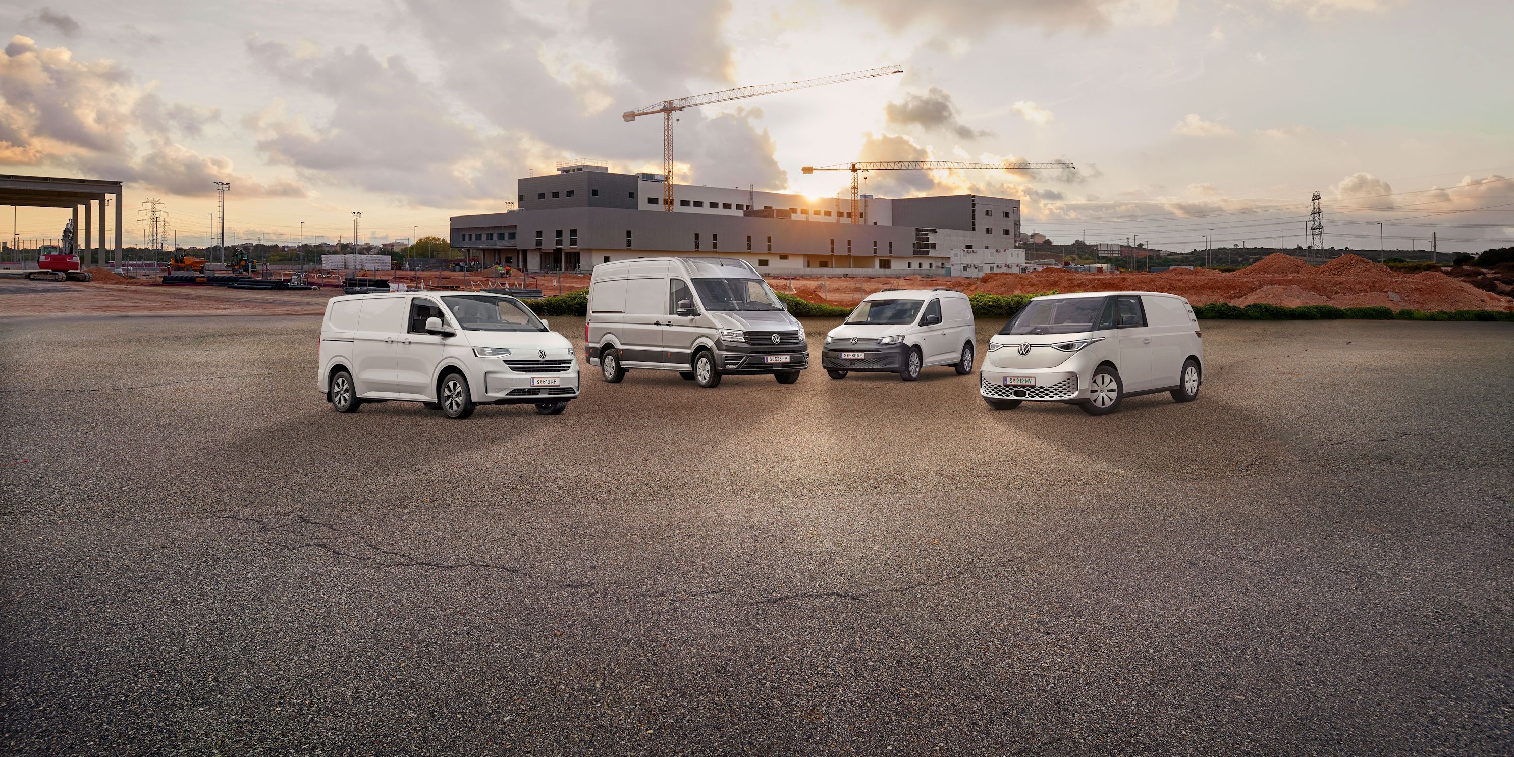 Vier weiße Transporter von Volkswagen Nutzfahrzeuge stehen nebeneinander auf einem leeren Parkplatz vor einem unfertigen Gebäude. Der Himmel ist bewölkt mit einigen Sonnenstrahlen. Ein Baukran ist im Hintergrund sichtbar.