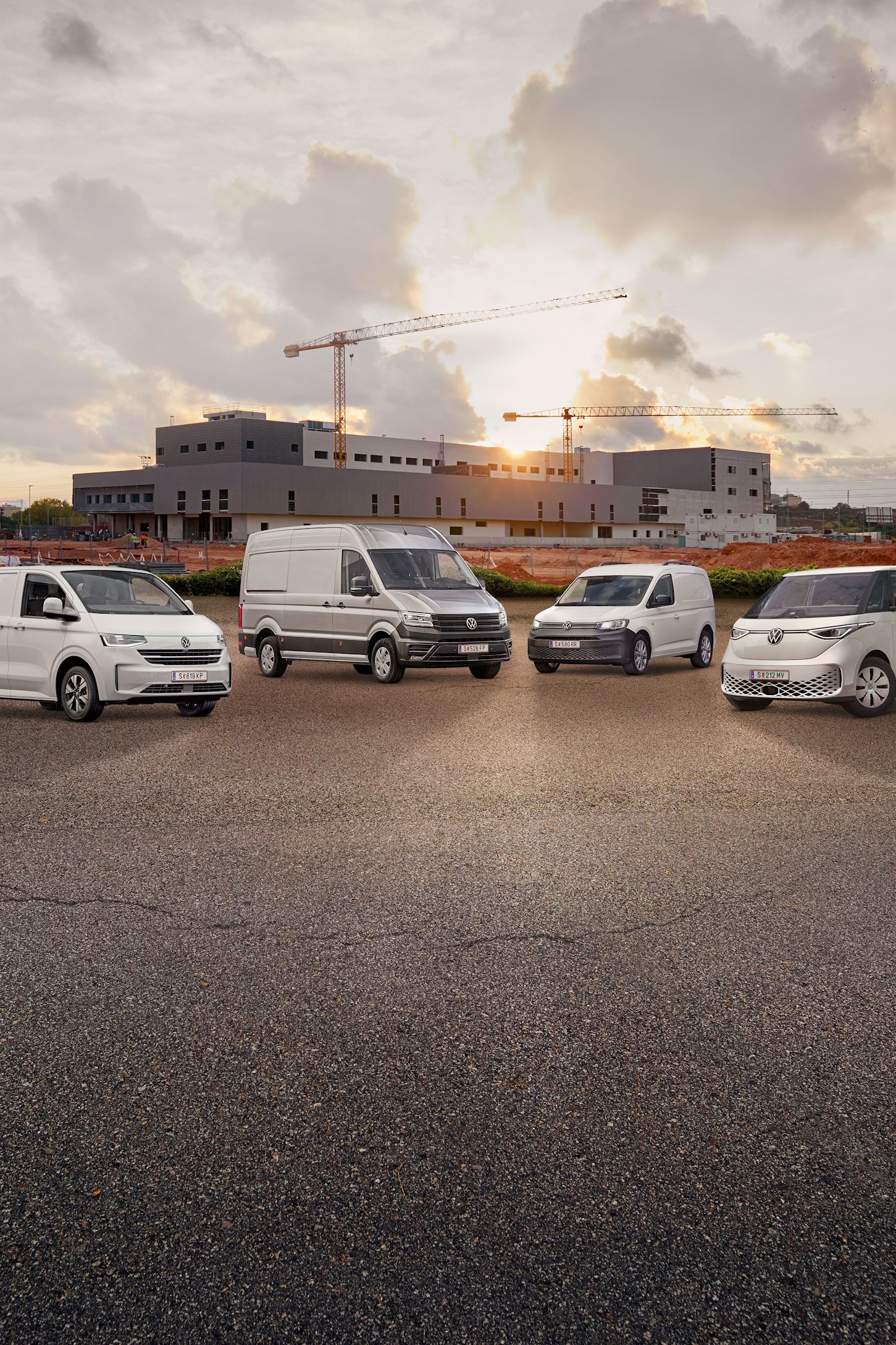 Vier weiße Transporter von Volkswagen Nutzfahrzeuge stehen nebeneinander auf einem leeren Parkplatz vor einem unfertigen Gebäude. Der Himmel ist bewölkt mit einigen Sonnenstrahlen. Ein Baukran ist im Hintergrund sichtbar.