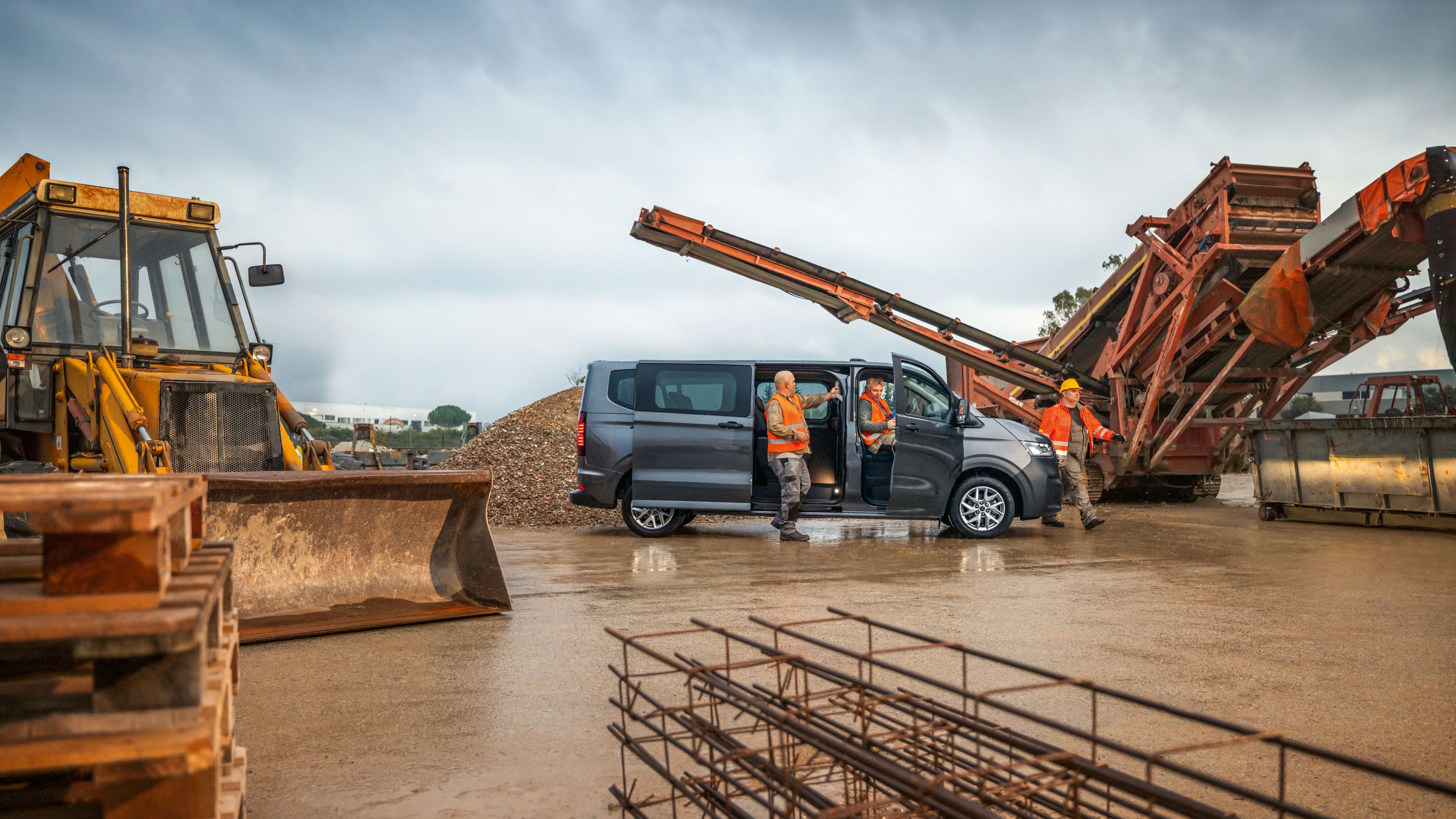 Bauarbeiter rund um einen grauen Volkswagen Transporter auf einer Baustelle.