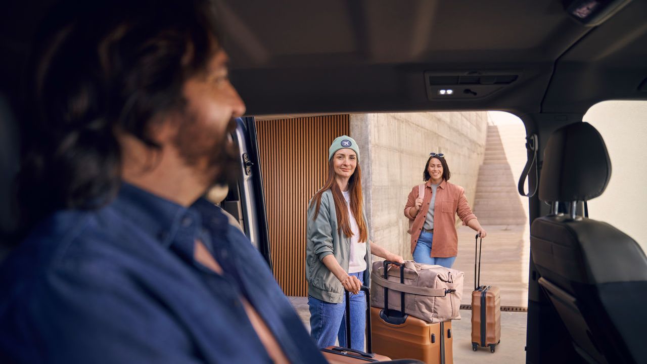 Zwei Personen stehen außerhalb eines Volkswagen Nutzfahrzeugs mit Gepäck, während eine Person im Vordergrund im Auto sitzt.