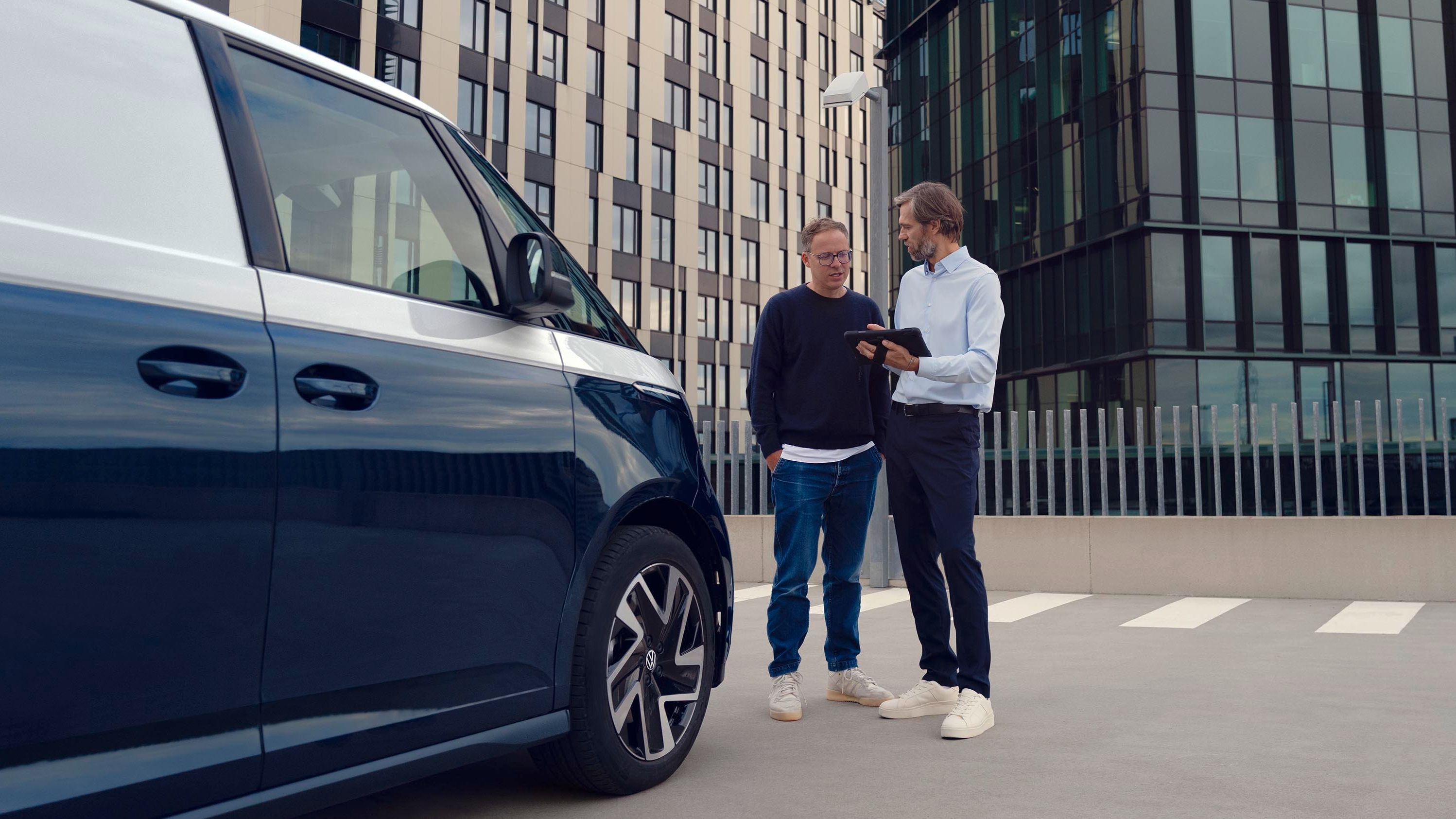 Zwei Personen stehen neben einem blauen Lieferwagen auf einem Parkplatz, umgeben von modernen Bürogebäuden. Eine Person zeigt der anderen etwas auf einem Tablet.