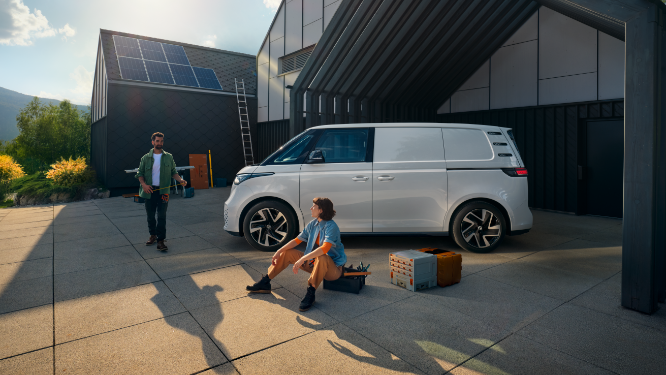 Ein weißer Volkswagen ID. Buzz Cargo vor einer Werkshalle, im Vordergrund sind ein Mann und eine Frau zu sehen