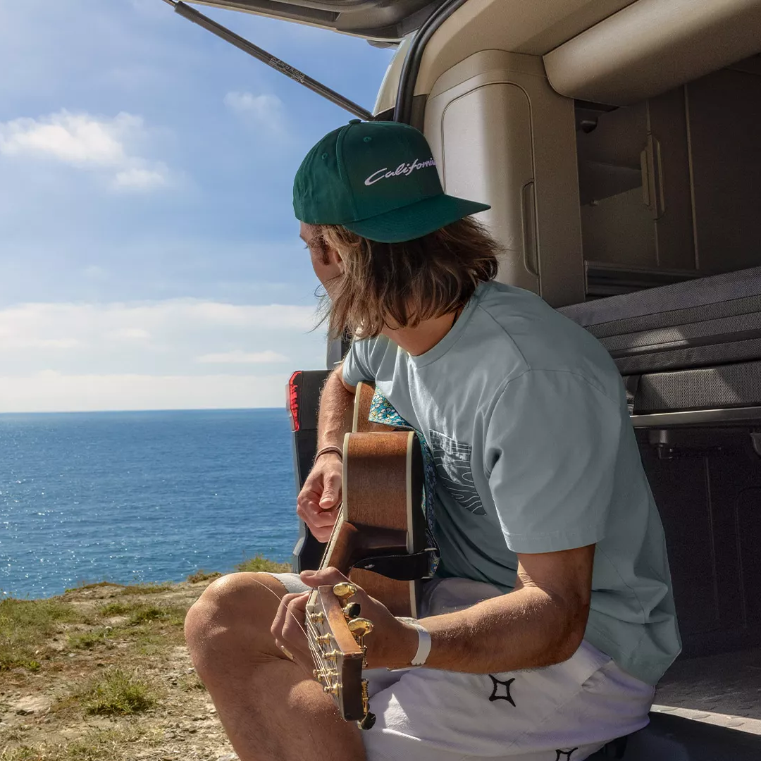 Gitarrenspielender Mann sitzt im geöffneten Kofferraum eines VW California und blickt auf das Meer. Er trägt ein grünes California Cap und ein hellblaues T-Shirt. Die Szene wirkt ruhig und idyllisch.