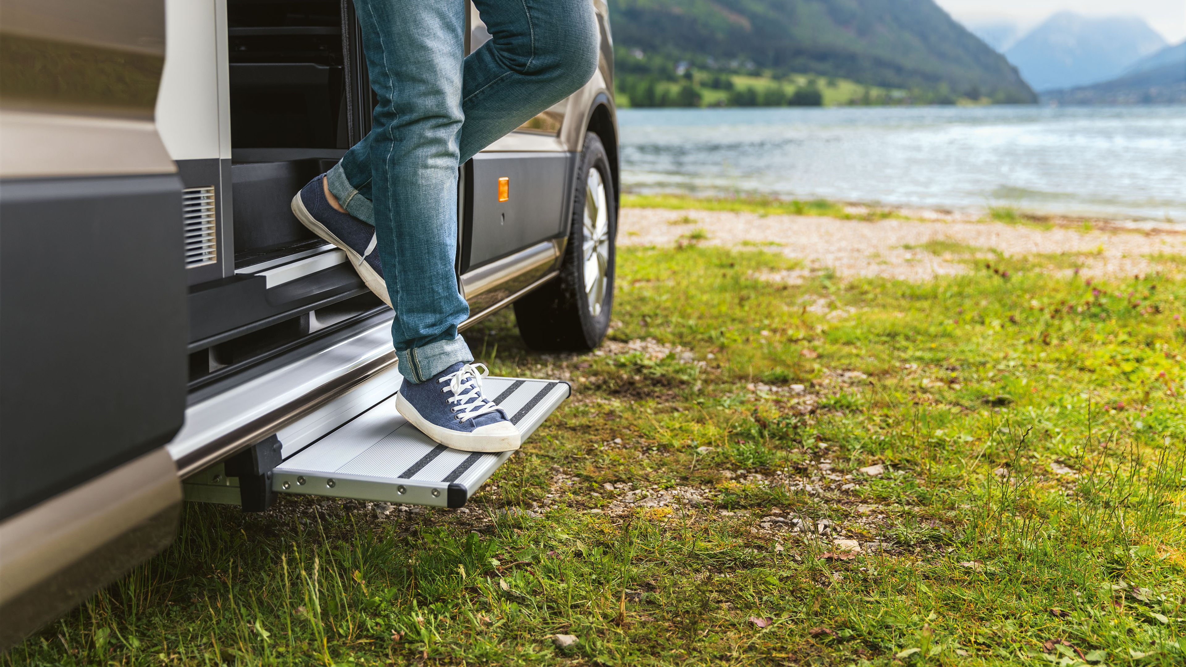 Großaufnahme der elektrisch ausfahrbaren Trittstufe vor einem Bergsee. Jemand steigt gerade entspannt aus. 