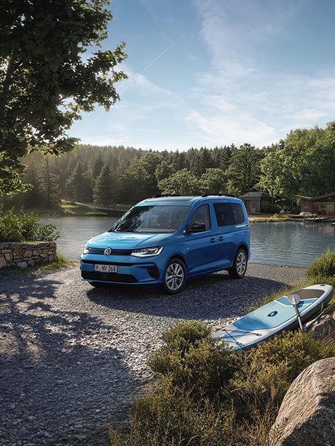 Ein blauer Volkswagen Caddy steht auf einem Schotterweg am Ufer eines ruhigen Sees, umgeben von dichtem, grünem Wald.