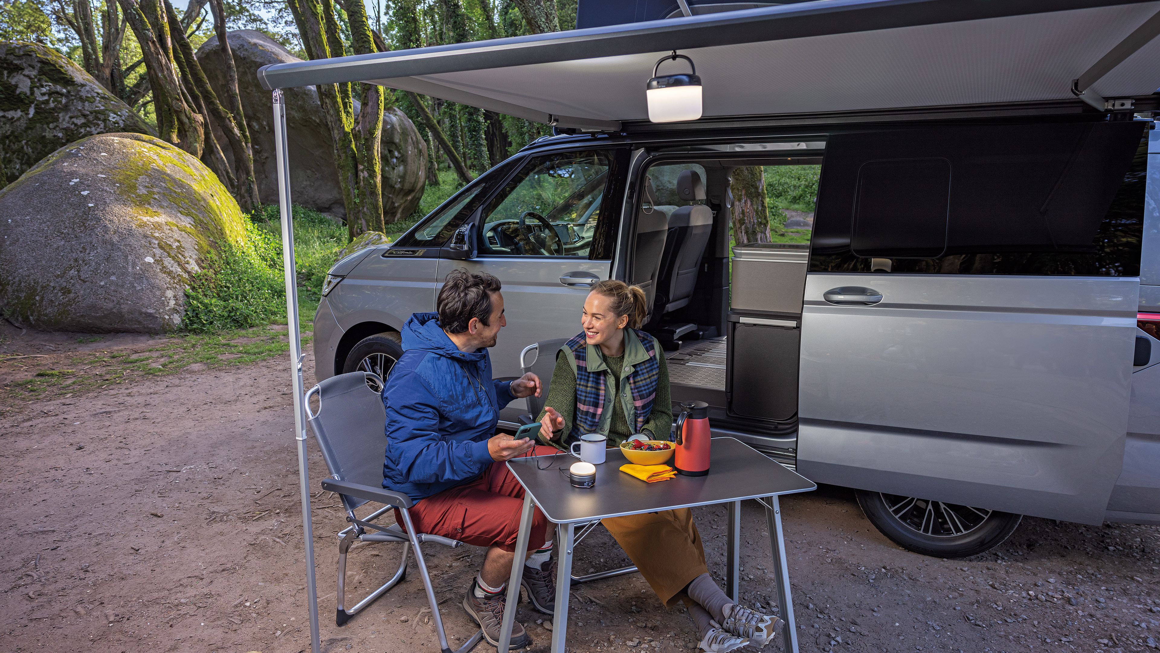 Ein Paar frühstückt unter der ausgefahrenen Marquise ihres New Californias, der im Wald parkt