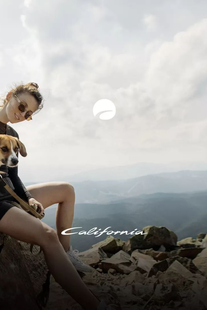 Eine Person sitzt auf einem felsigen Gipfel und hält einen Hund neben sich. Die Landschaft im Hintergrund zeigt hügelige Berge und bewölkten Himmel. Das Wort "California" ist am Bildrand zu sehen.
