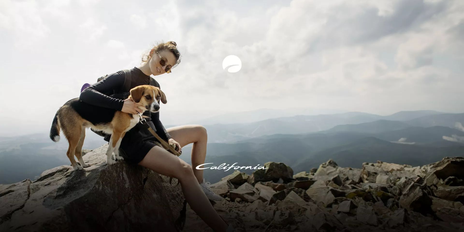 Eine Person sitzt auf einem felsigen Gipfel und hält einen Hund neben sich. Die Landschaft im Hintergrund zeigt hügelige Berge und bewölkten Himmel. Das Wort "California" ist am Bildrand zu sehen.