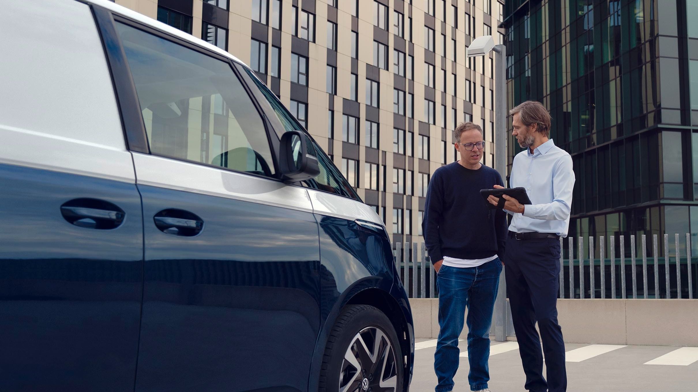 Zwei Personen stehen neben einem blauen Lieferwagen auf einem Parkplatz, umgeben von modernen Bürogebäuden. Eine Person zeigt der anderen etwas auf einem Tablet.