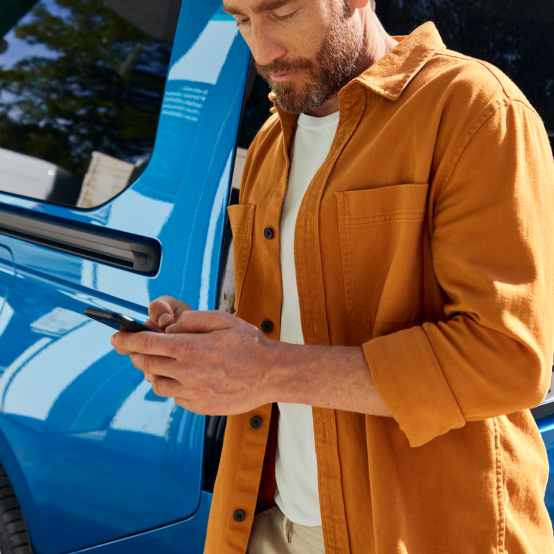 Ein Mann in einer senfgelben Jacke steht vor einem blauen Fahrzeug und schaut auf sein Smartphone.