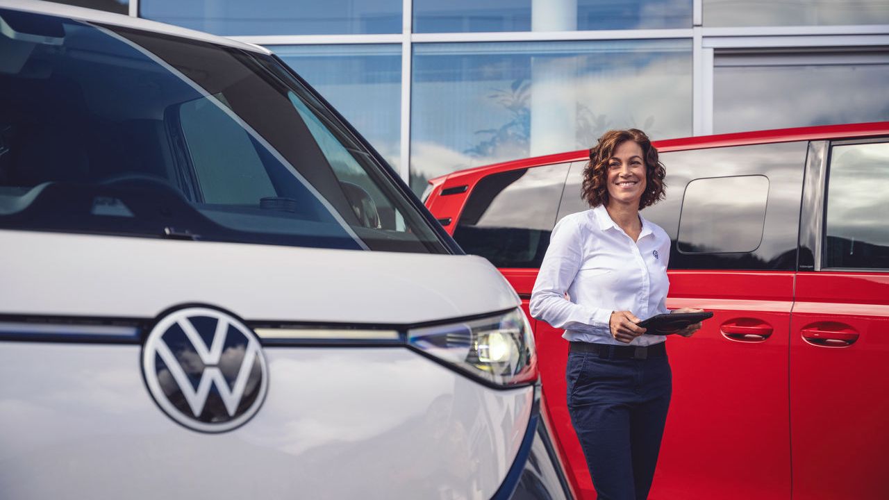 Eine Volkswagen Mitarbeiterin hält ein Tablet in ihren Händen. Links von Ihr steht ein blau weißer und rechts ein roter ID. Buzz.