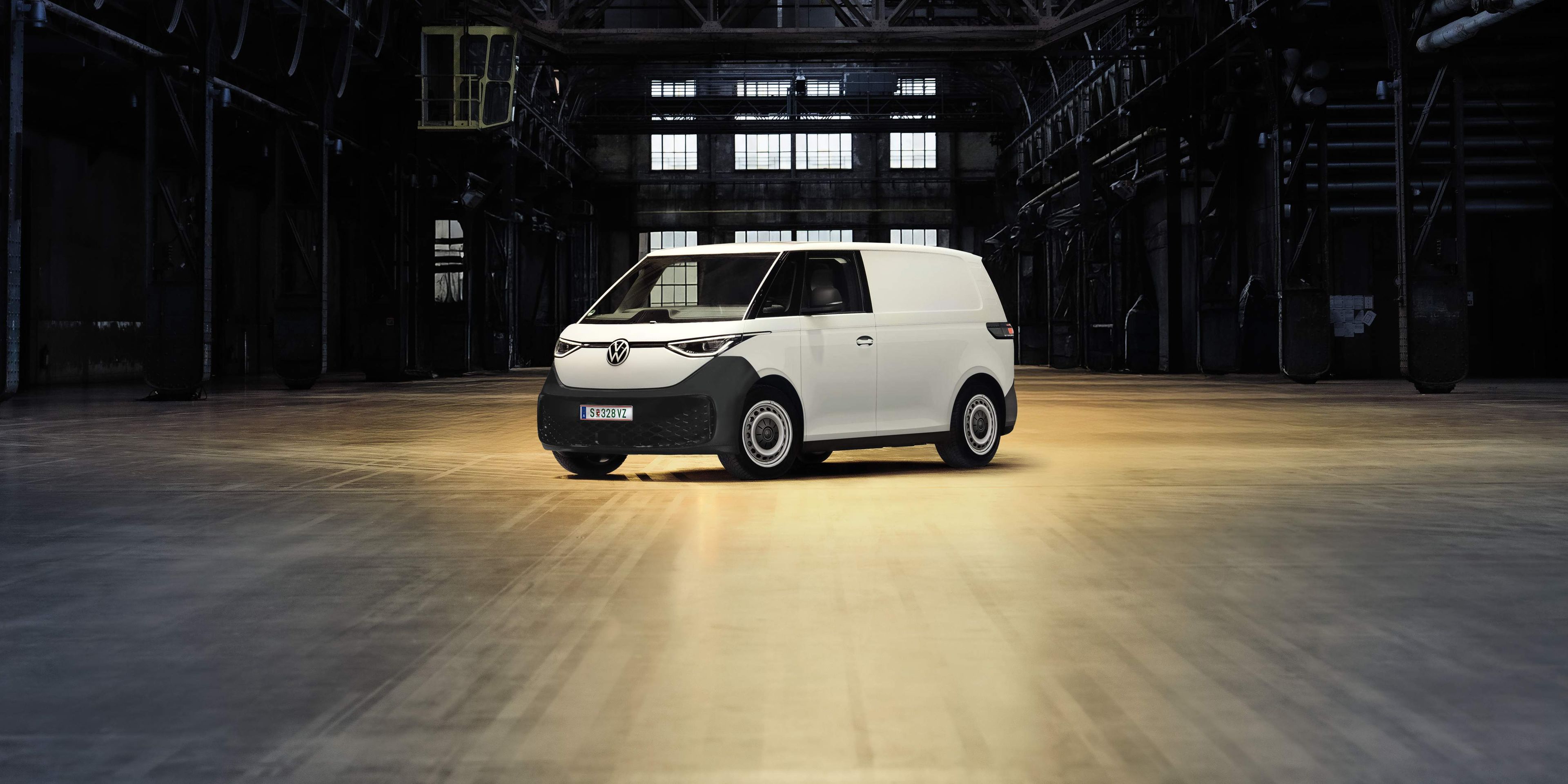Ein weißer Lieferwagen steht allein in einer großen, dunklen Industriehalle. Die Umgebung ist leer und weist Metallstrukturen sowie Fenster im Hintergrund auf. Ein einzelner Scheinwerfer beleuchtet den Lieferwagen.