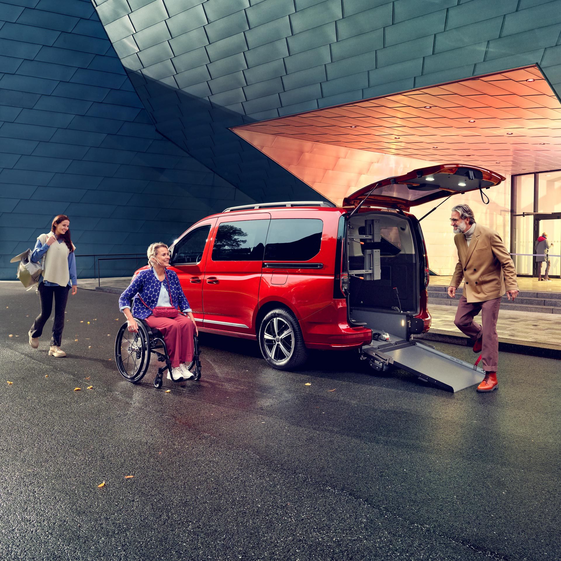 Ein roter VW Caddy mit offener Heckklappe und Rampe, eine Frau im Rollstuhl fährt darauf zu