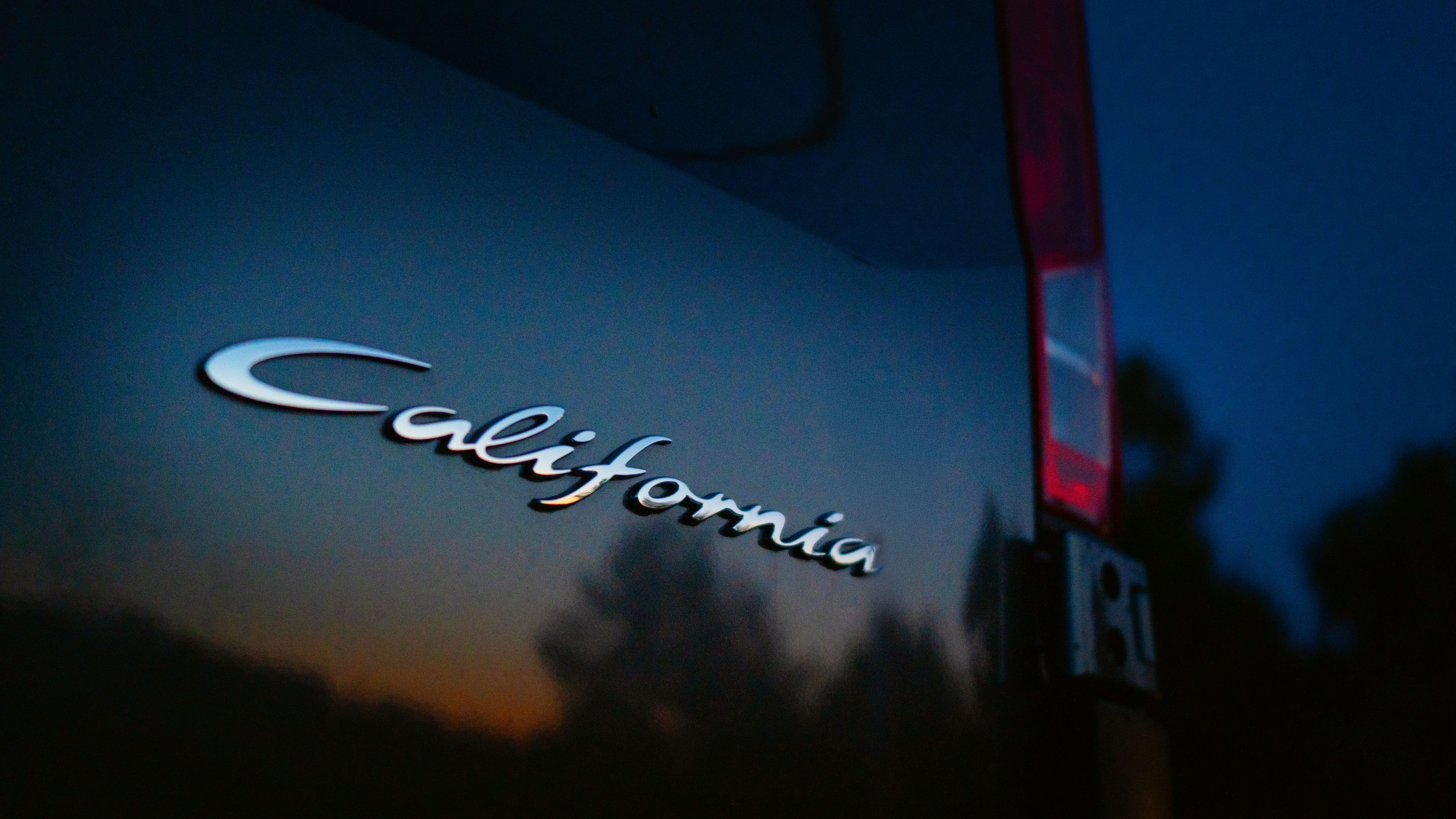 Nahaufnahme eines Autos bei Sonnenuntergang, das Wort „California“ ist auf der Seite des Fahrzeugs geschrieben. Die Umgebung ist dunkel, und die Silhouette eines Baumes ist sichtbar.