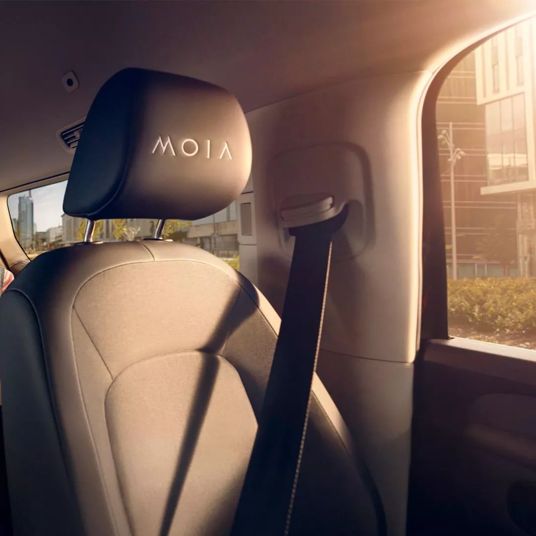 Ein Kind sitzt auf dem Rücksitz eines Autos und spielt auf einem Tablet, während ein Erwachsener neben ihm sitzt. Die Sitze haben einen schwarzen Bezug mit dem Logo "MOIA". Das Auto ist geräumig und von Tageslicht durchflutet.