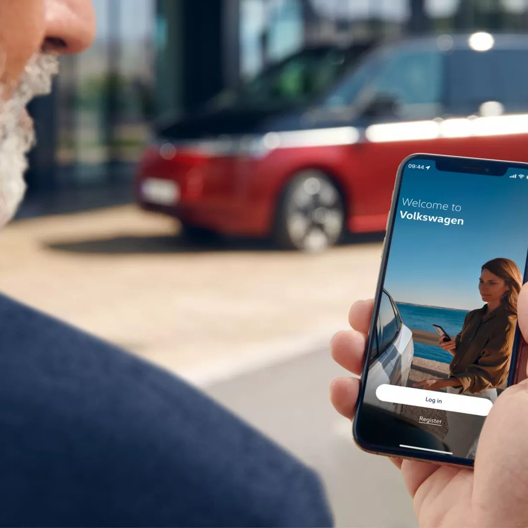 Eine Person hält ein Smartphone, auf dessen Bildschirm eine Willkommensnachricht von Volkswagen angezeigt wird. Im Hintergrund ist ein rotes Auto zu sehen.