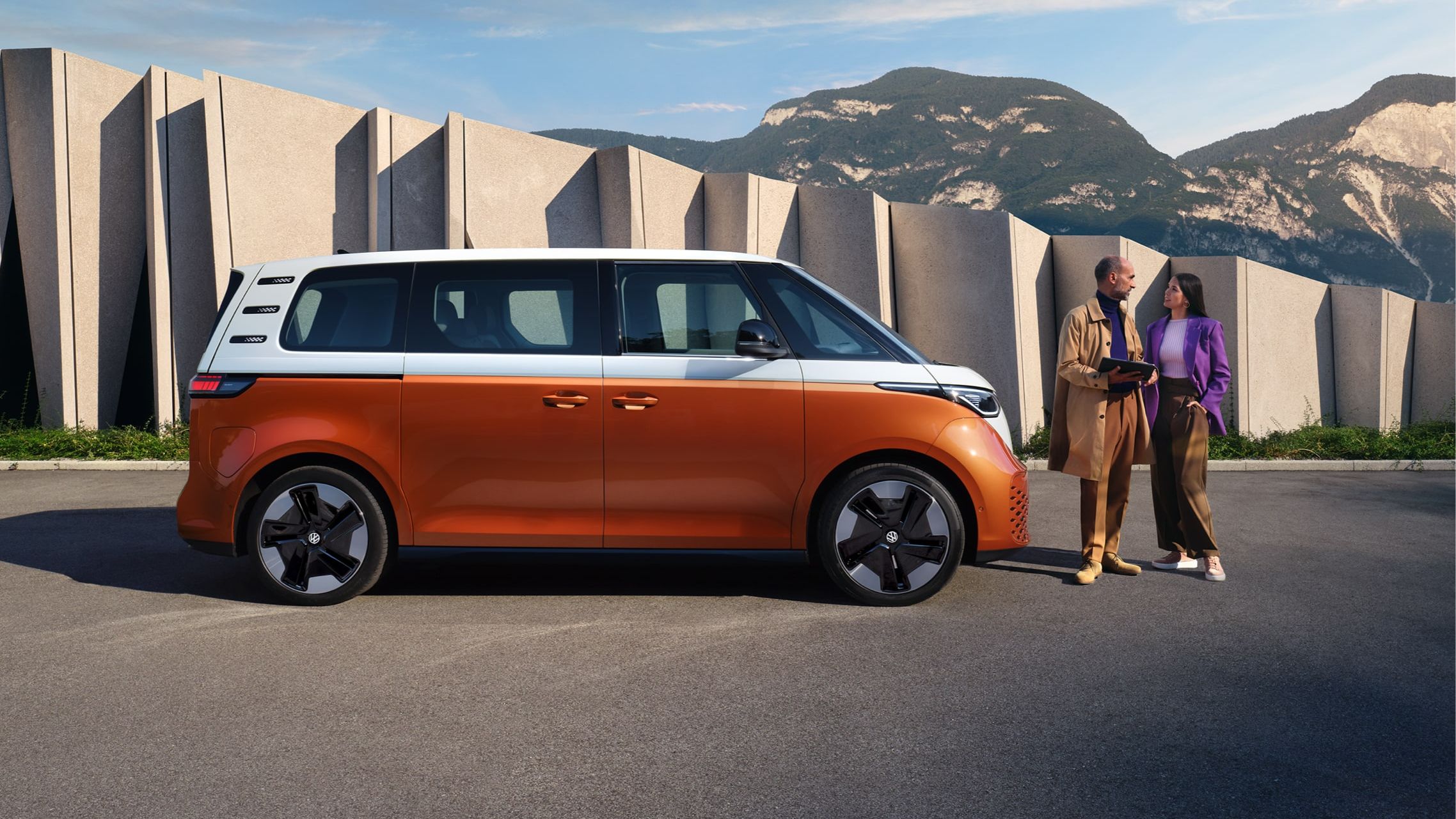 Zwei Personen unterhalten sich vor einem orange weißer ID. Buzz mit Gebäude und Bergen im Hintergrund 
