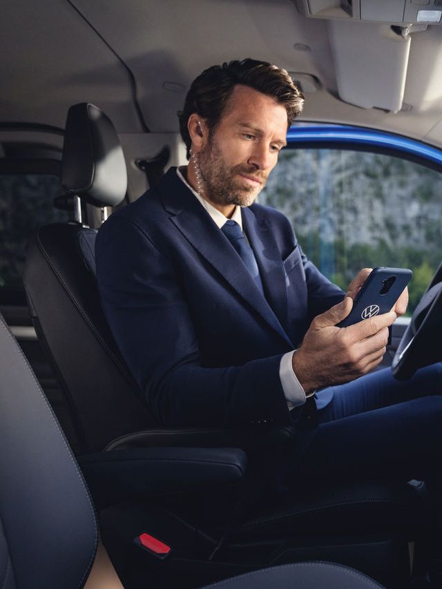 Ein Mann im Anzug sitzt auf dem Fahrersitz eines Volkswagen Nutzfahrzeuges und blickt konzentriert auf sein Smartphone in einer Hülle mit Volkswagen Logo.