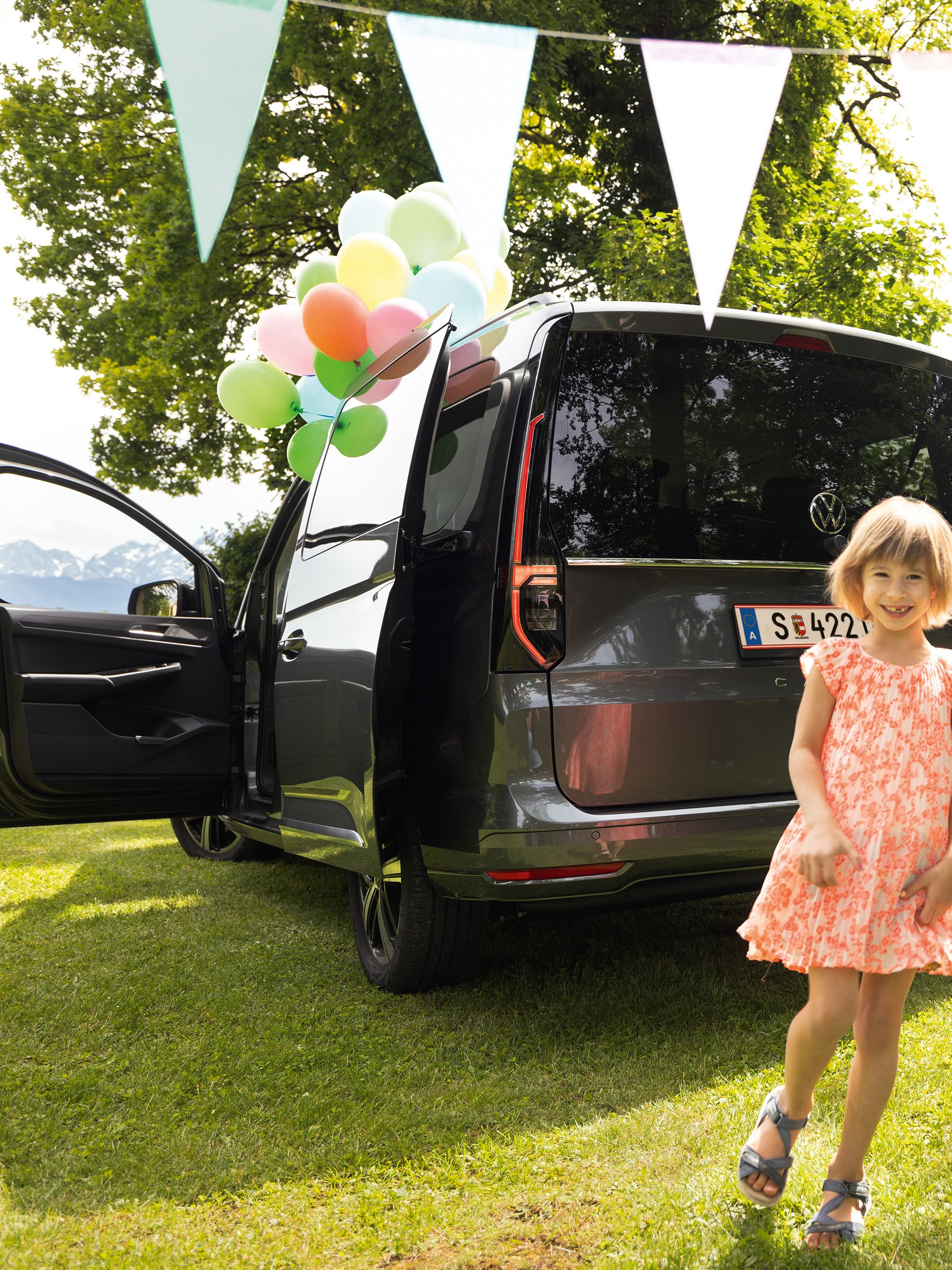 VW Caddy steht im Garten und ist mit Ballons geschmückt, Kinder spielen neben dem Fahrzeug