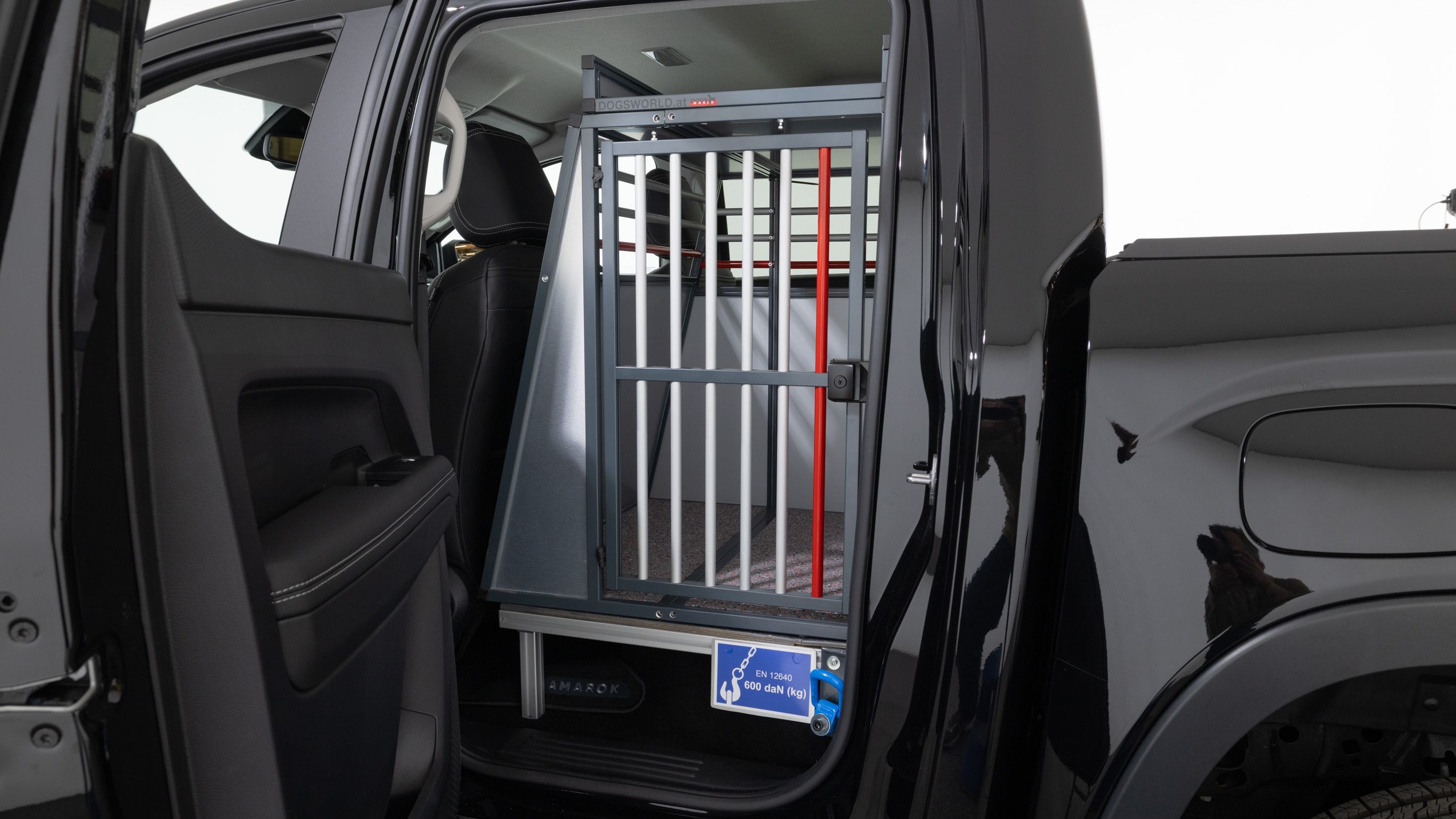 Eine Rückbank eines schwarzen Autos wurde ausgebaut und durch einen Tierkäfig ersetzt. Der Käfig befindet sich direkt hinter dem Fahrersitz und ist durch eine Tür mit Gitterstäben zugänglich. Außen ist ein Schild sichtbar, auf dem ein Hund abgebildet ist.