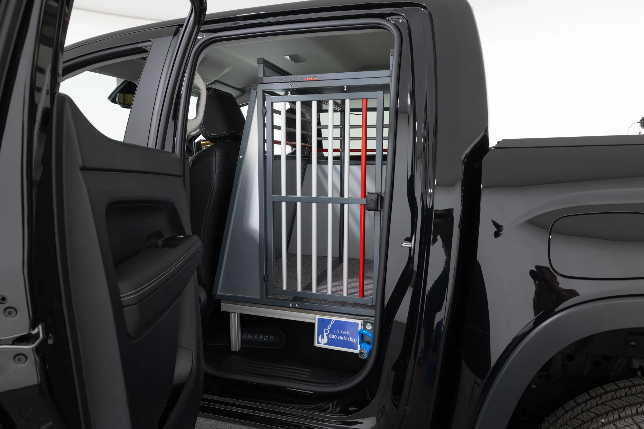 Eine Rückbank eines schwarzen Autos wurde ausgebaut und durch einen Tierkäfig ersetzt. Der Käfig befindet sich direkt hinter dem Fahrersitz und ist durch eine Tür mit Gitterstäben zugänglich. Außen ist ein Schild sichtbar, auf dem ein Hund abgebildet ist.