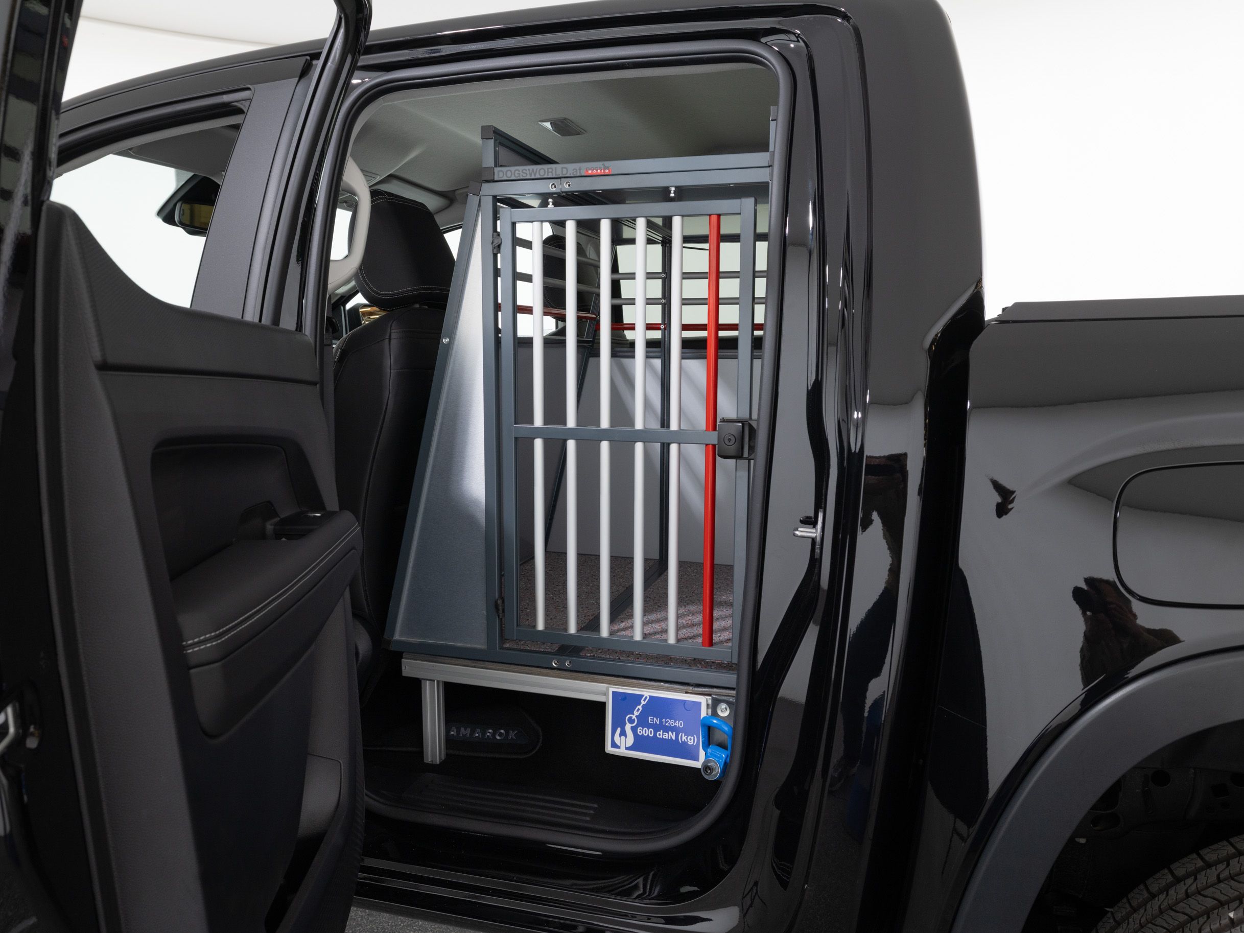 Eine Rückbank eines schwarzen Autos wurde ausgebaut und durch einen Tierkäfig ersetzt. Der Käfig befindet sich direkt hinter dem Fahrersitz und ist durch eine Tür mit Gitterstäben zugänglich. Außen ist ein Schild sichtbar, auf dem ein Hund abgebildet ist.